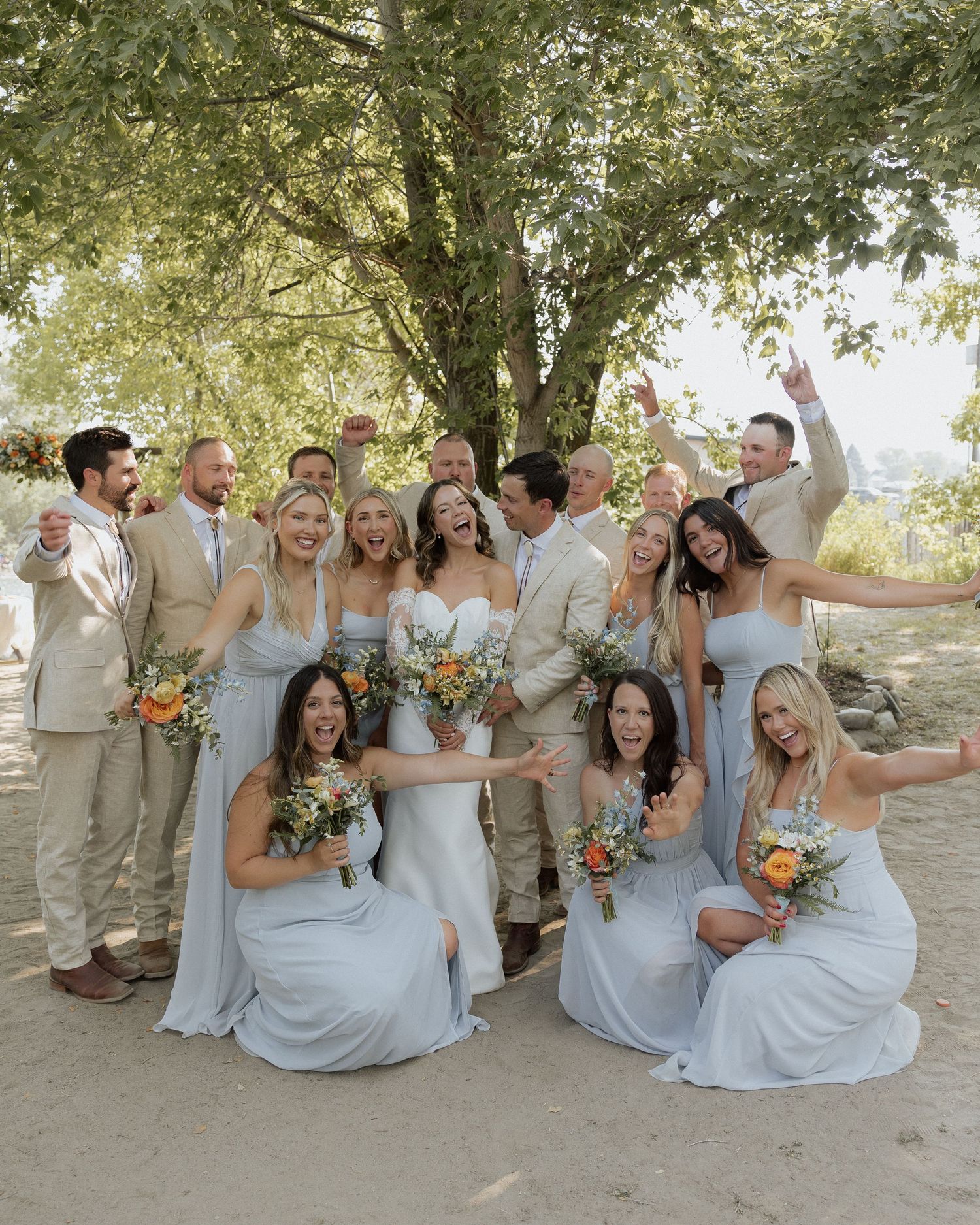 Joyful wedding party in pale blue dresses and beige suits celebrating with fun poses outdoors.