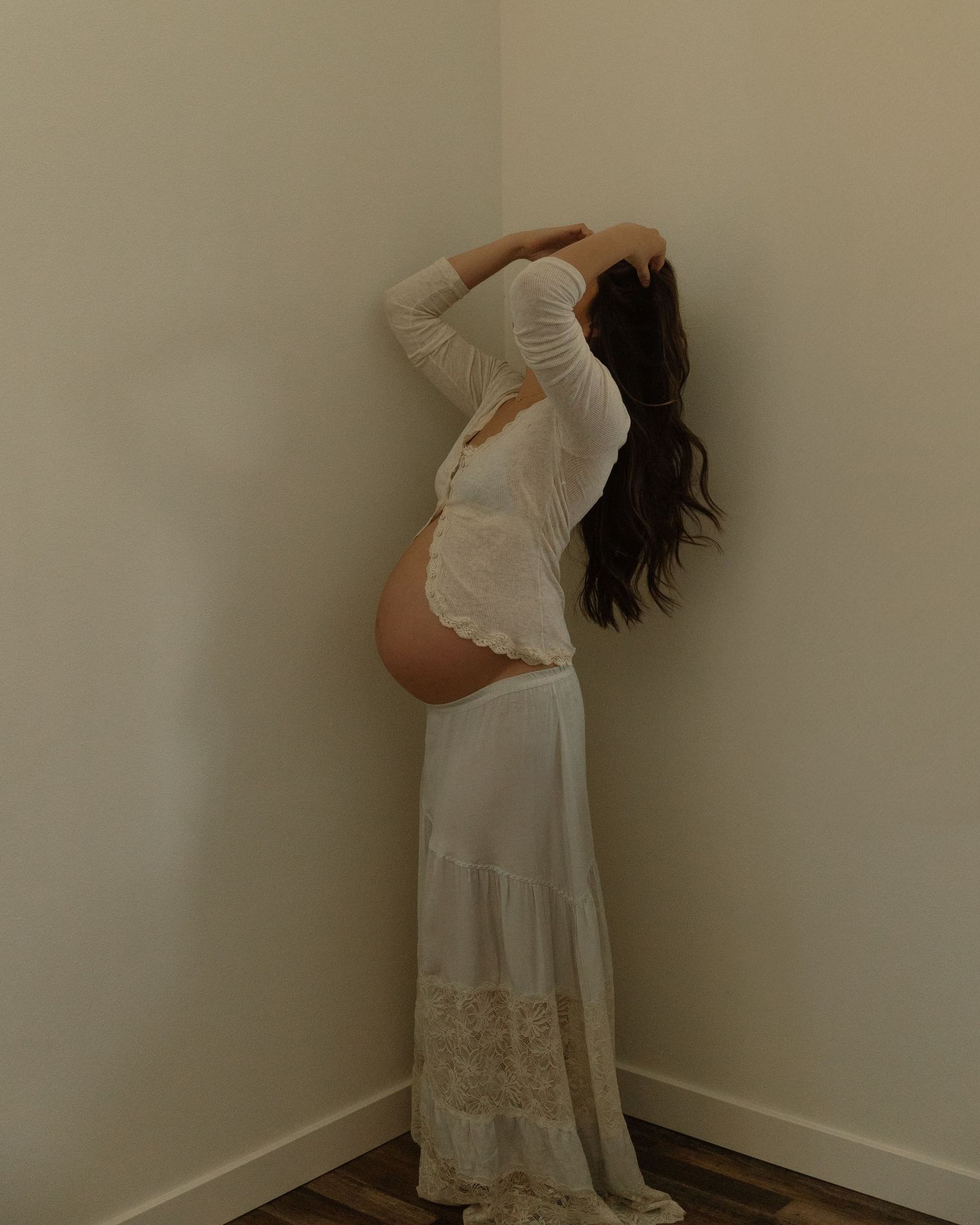 A side view of an expectant mother in a flowing white lace dress against a beige wall.
