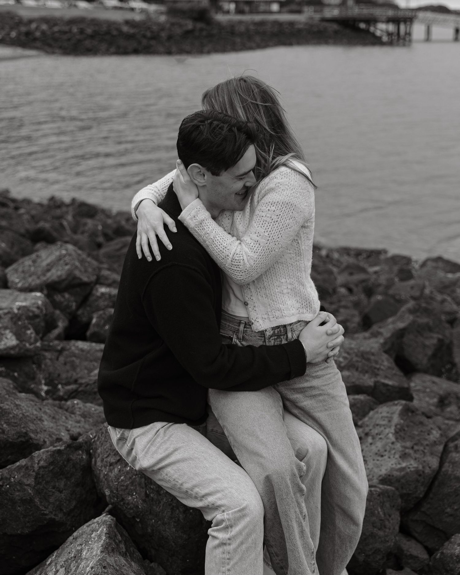 Two people in a loving embrace seated on coastal rocks by the water.
