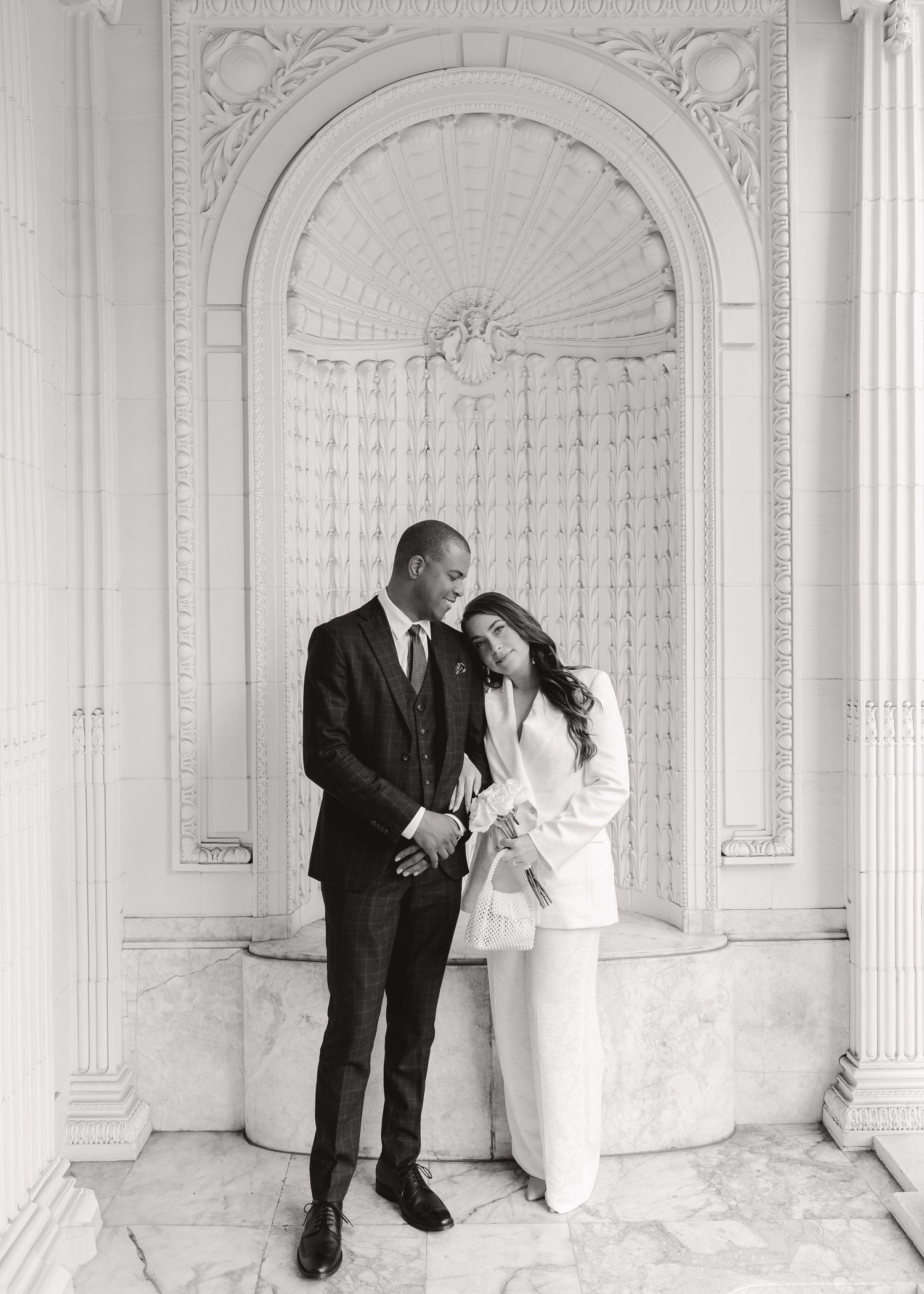 Black and white photo of an elegant couple in formal attire standing before an ornate marble arch.
