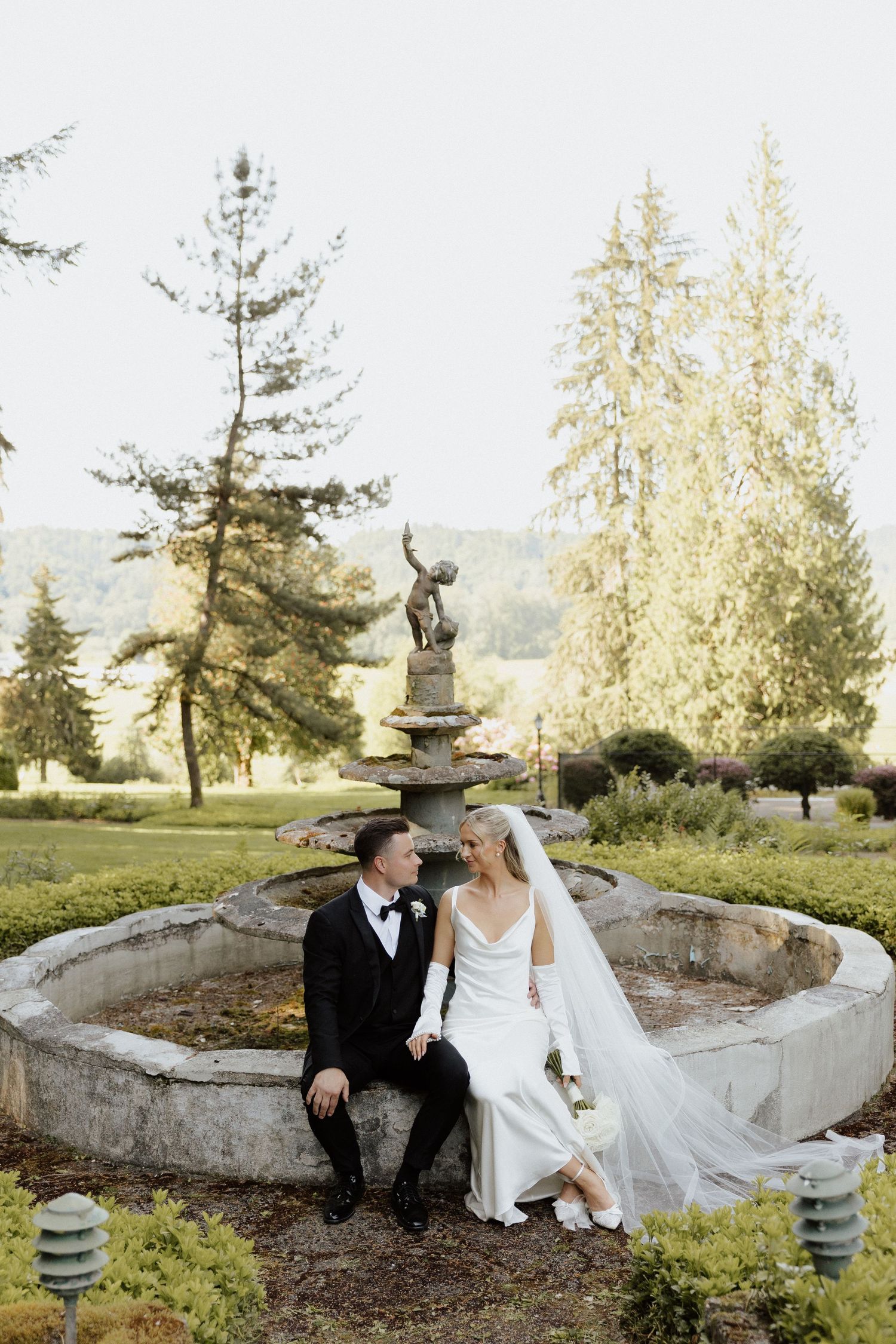 Bride and groom sitting together at stone fountain in lush garden on their wedding day.