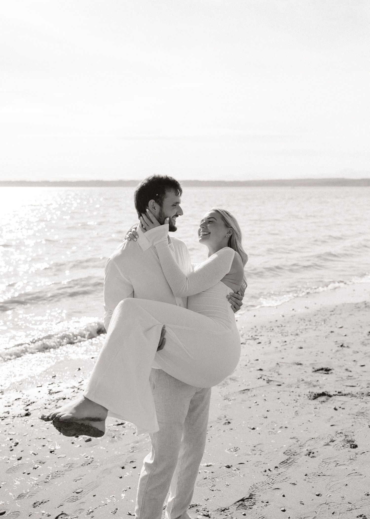 Black and white beach wedding photo of groom carrying bride in white dress, laughing together by ocean waves.