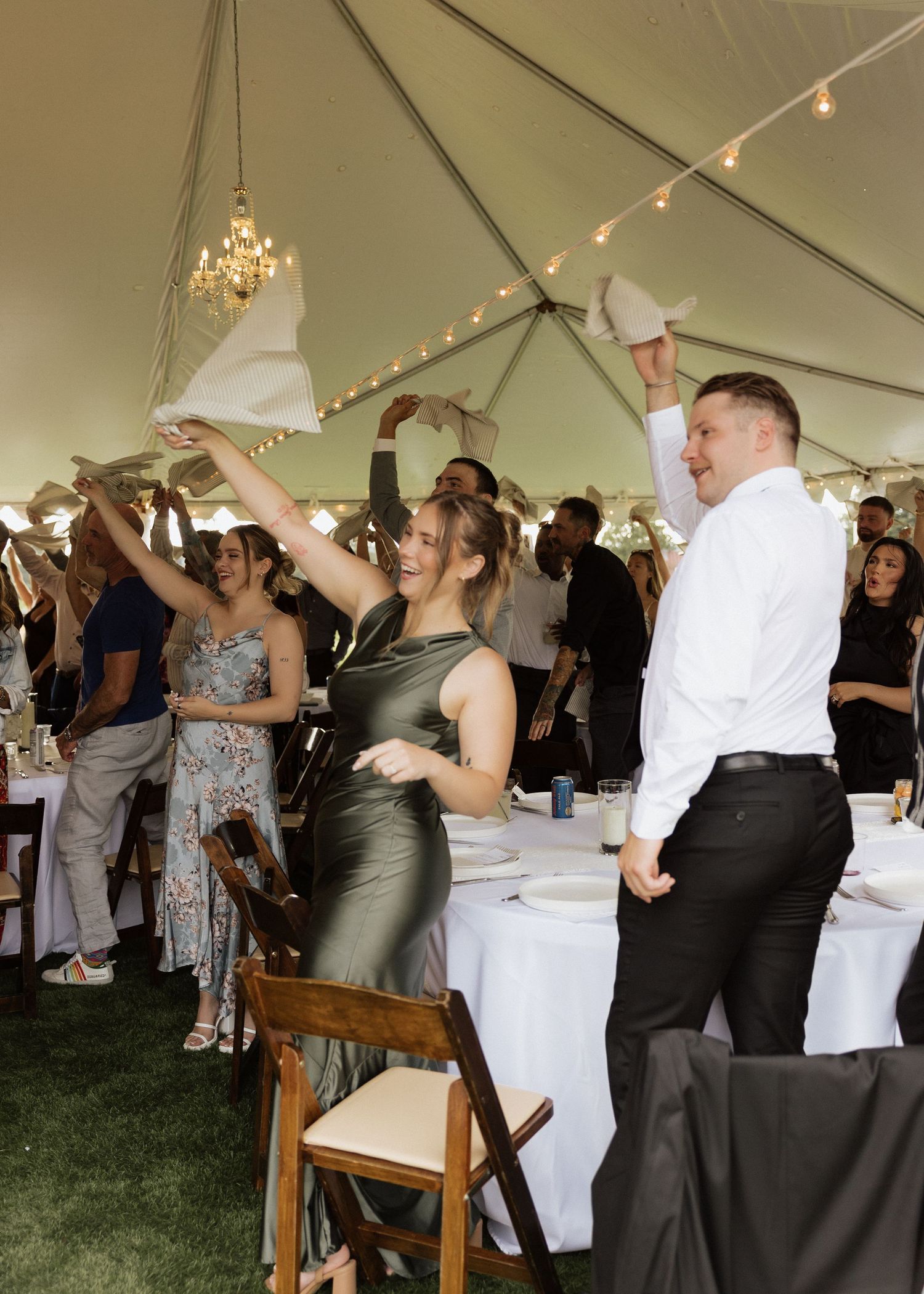 Guests wave napkins and celebrate at an elegant outdoor wedding reception under a tent with string lights.