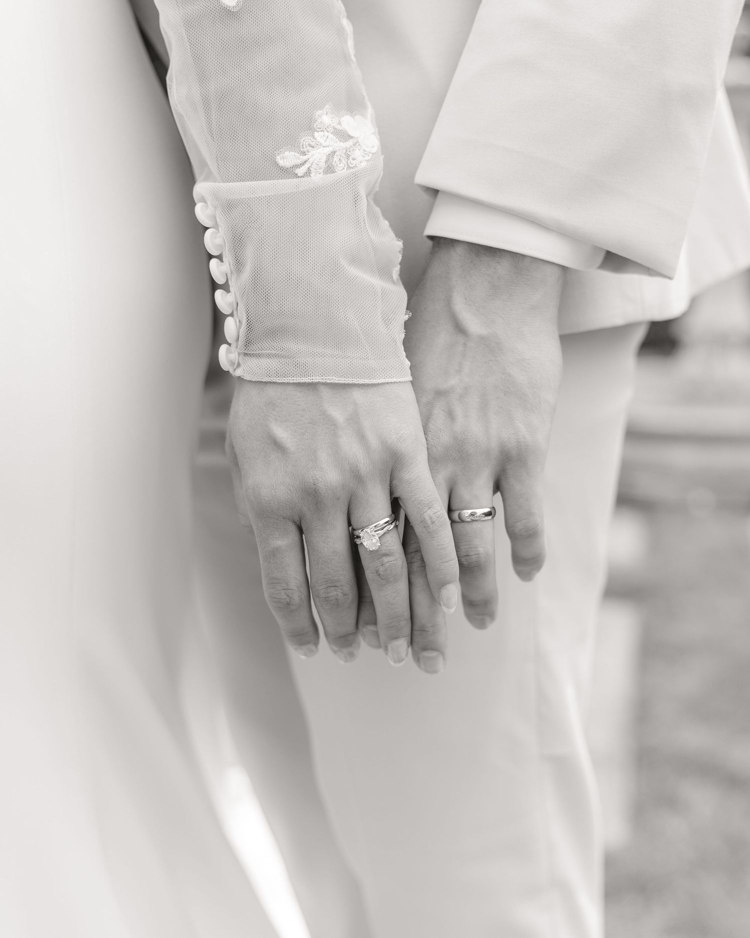 Black and white photo of newlyweds hands showing wedding rings, bride wearing lace sleeve gown beside groom in suit.