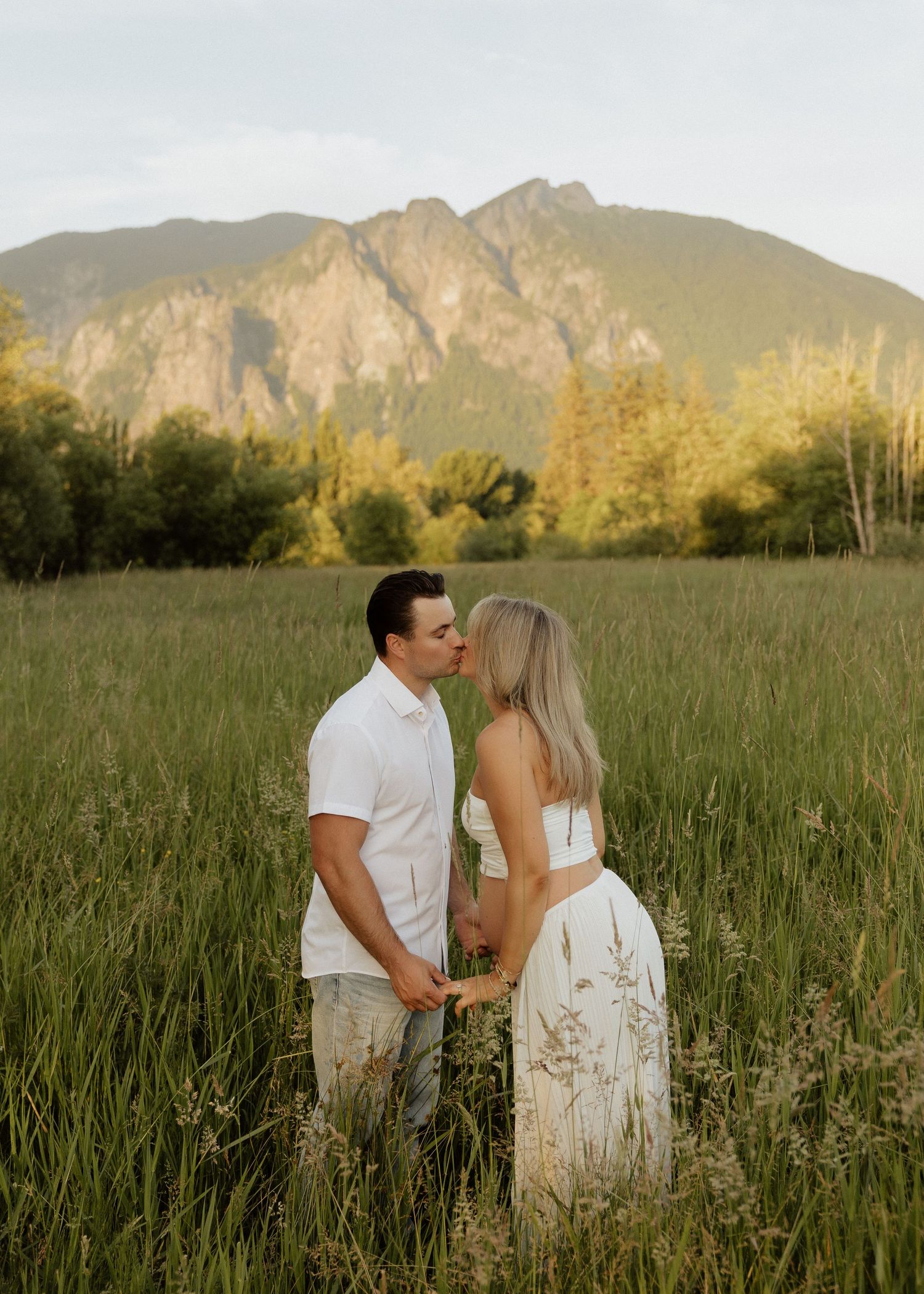 Couple kissing in a tall grass field with a dramatic mountain range visible in the golden hour background.