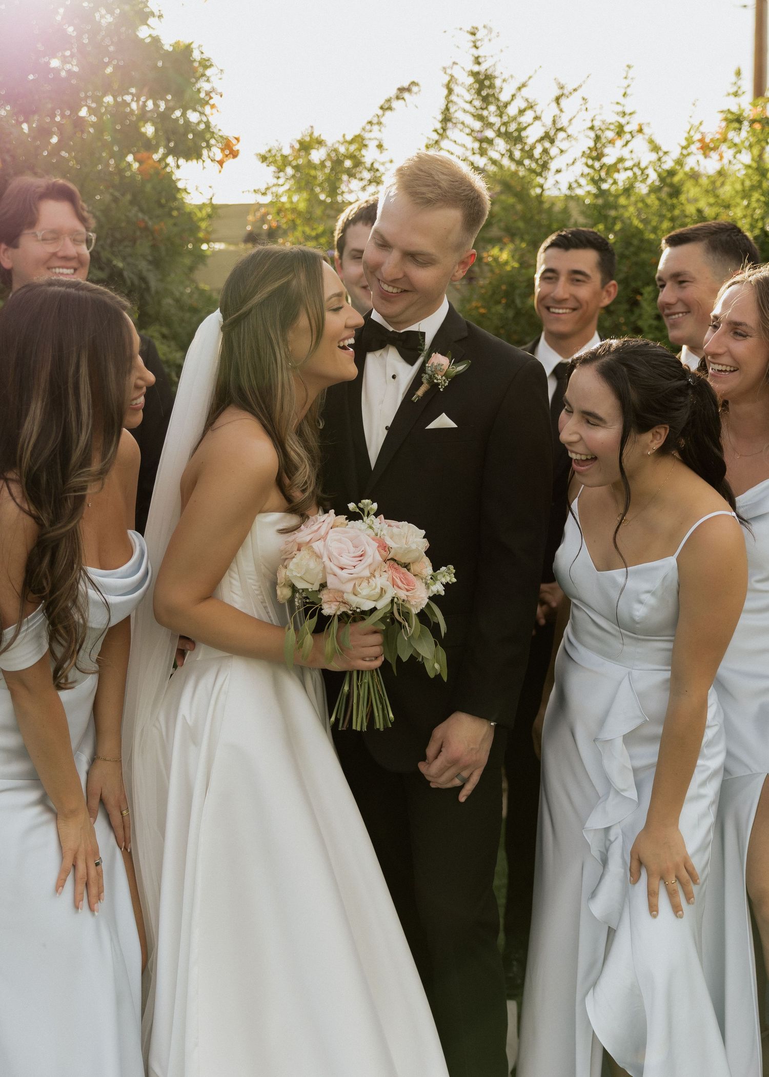 Bride and groom share a joyful moment surrounded by smiling wedding party guests in formal attire.