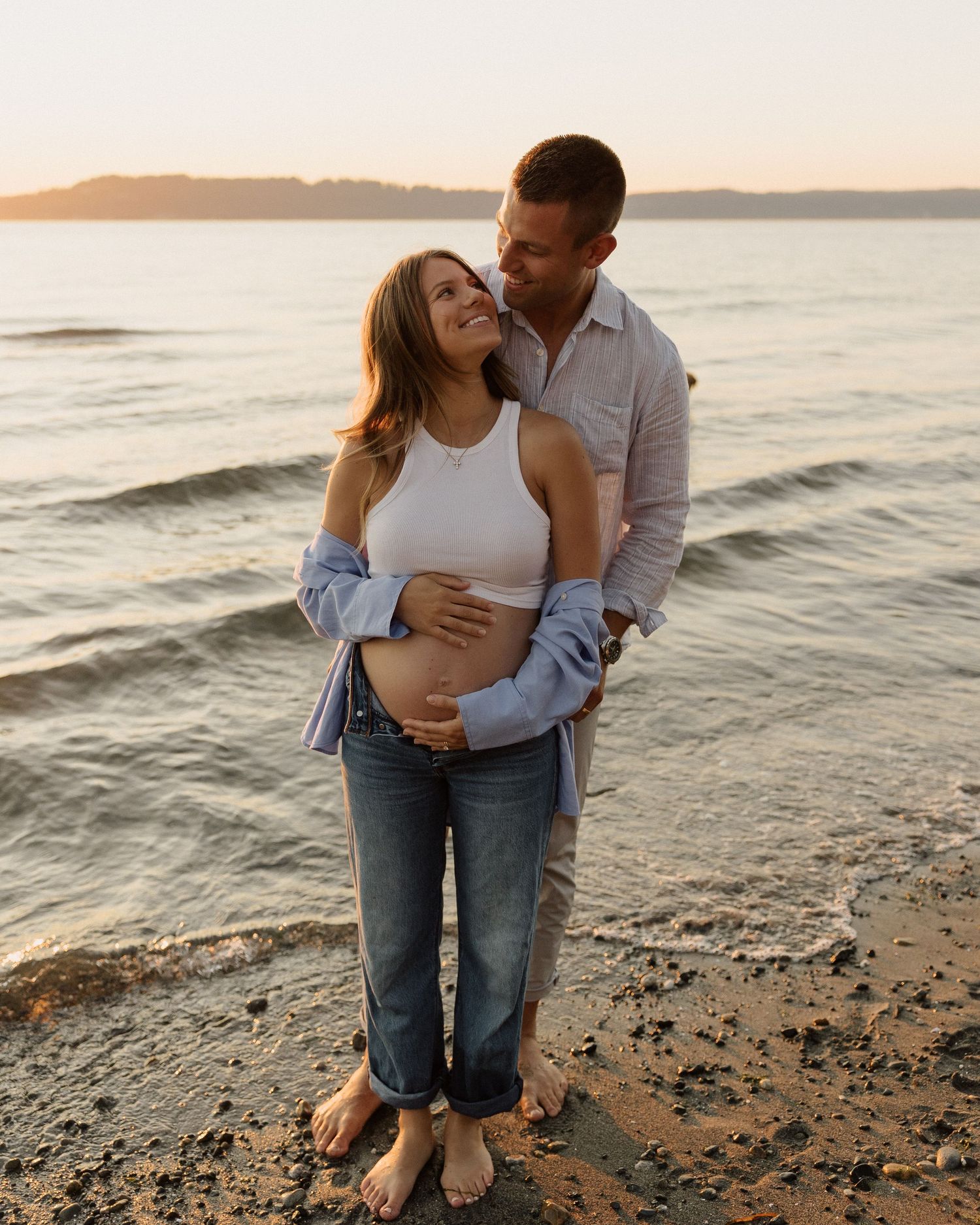 A loving couple poses for a maternity photo on a rocky beach during sunset.