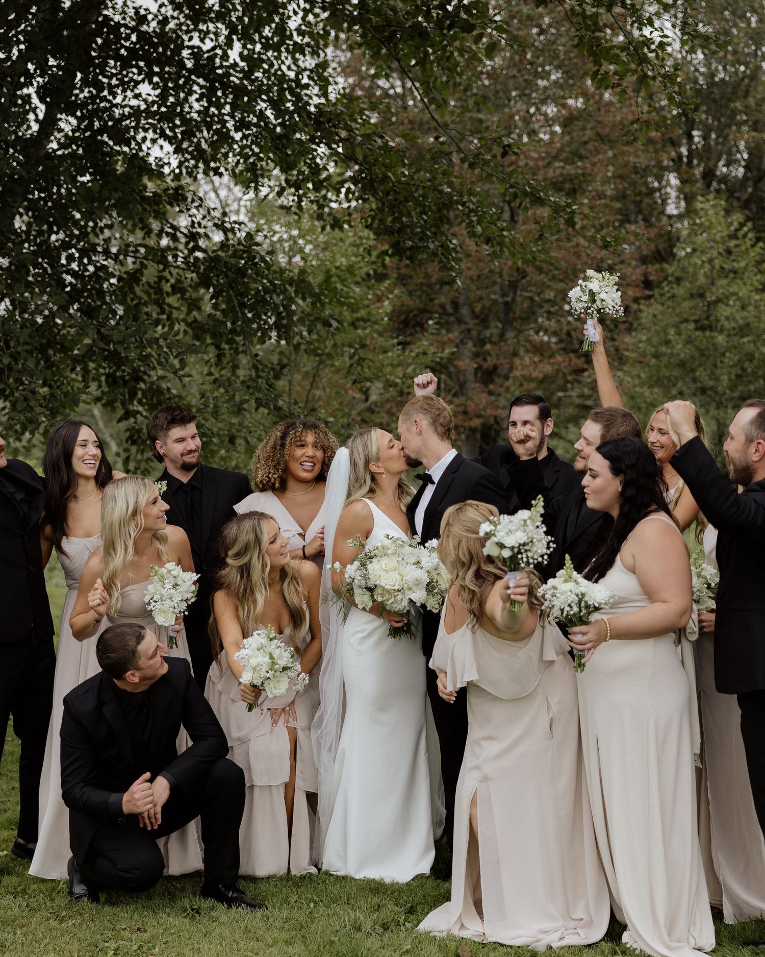 Wedding party in blush and black attire gathers for group photo in garden setting.
