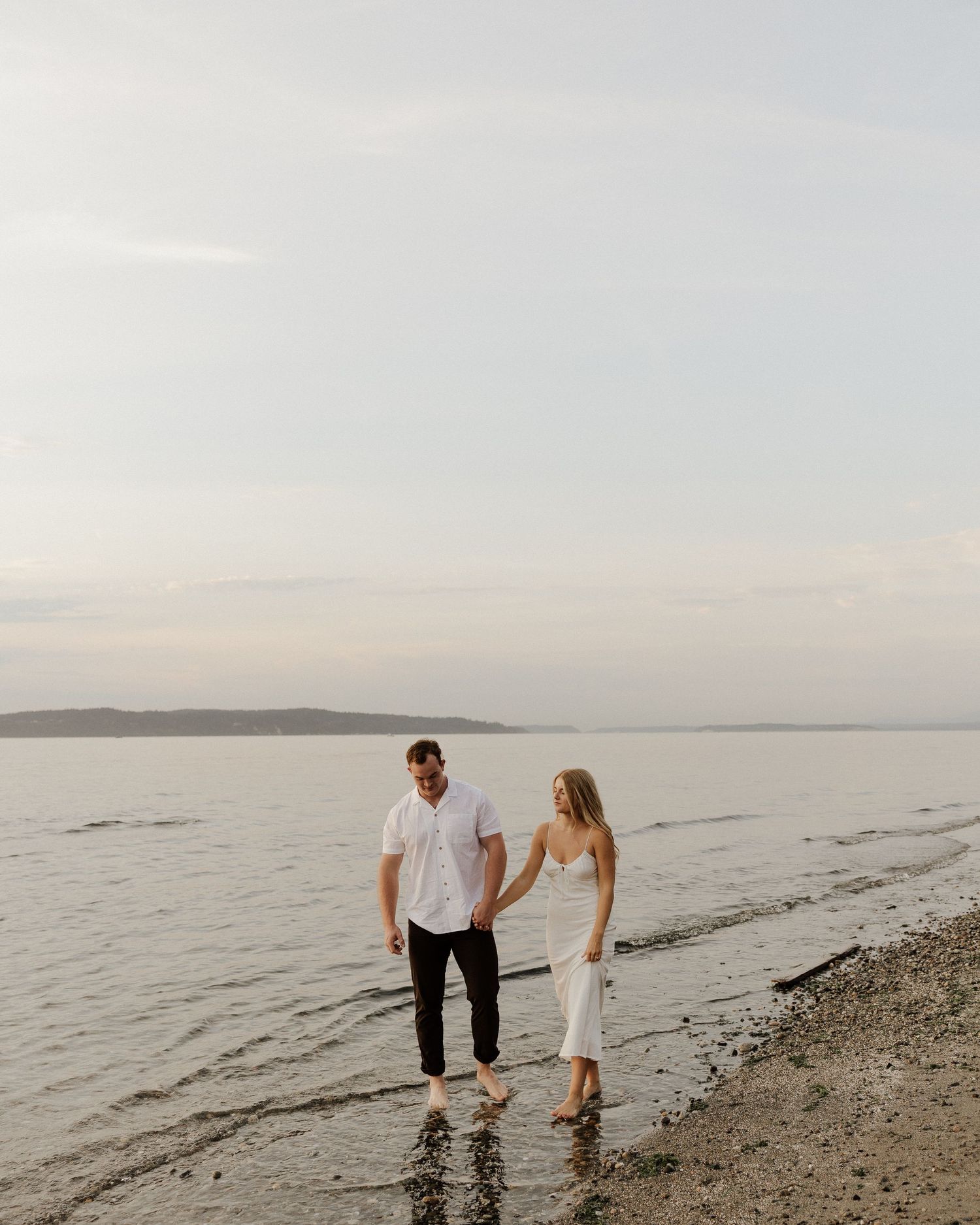 A couple walks barefoot together on a rocky beach during golden hour.