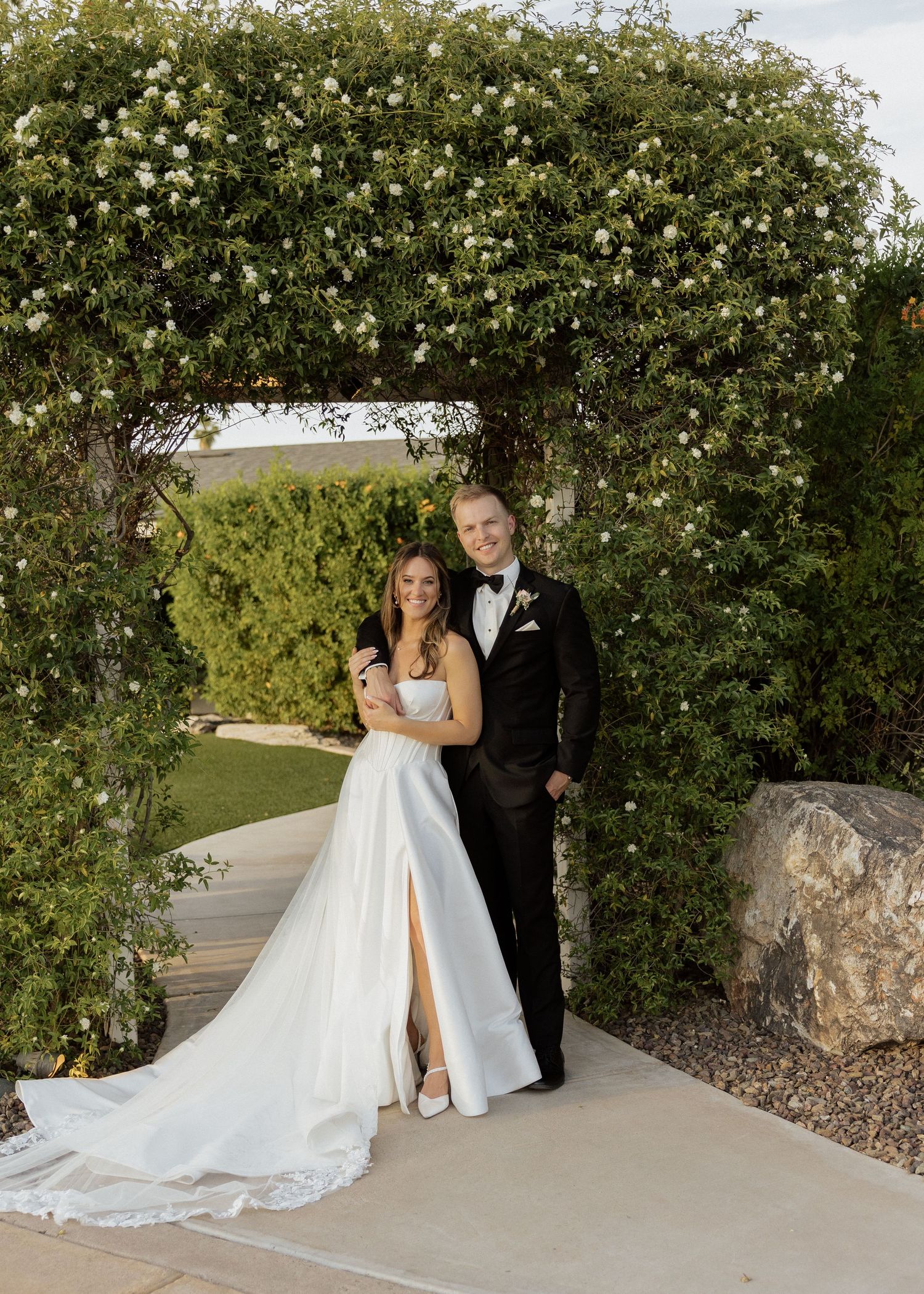 Bride in white gown and groom in black tuxedo pose under floral arch at outdoor wedding venue.