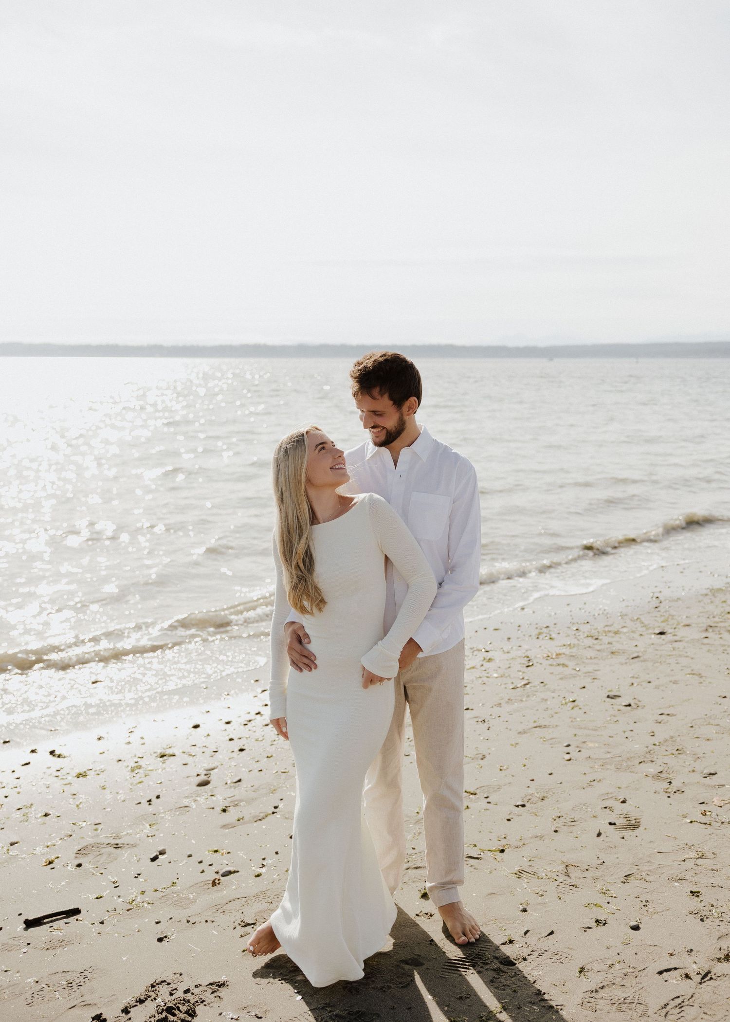 A couple in white outfits share a romantic moment on a sandy beach with calm ocean waters behind them.