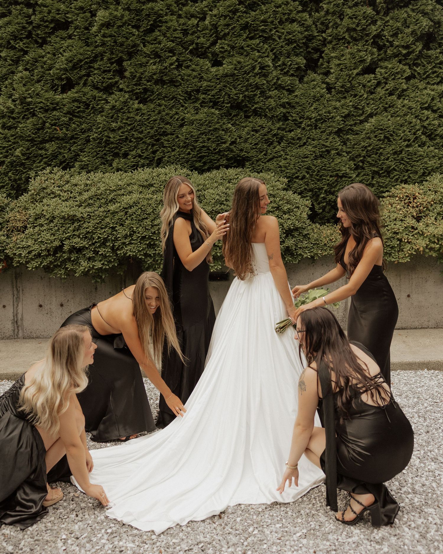 Bridesmaids in black dresses arrange the train of a bride in a white gown outdoors against a green hedge.