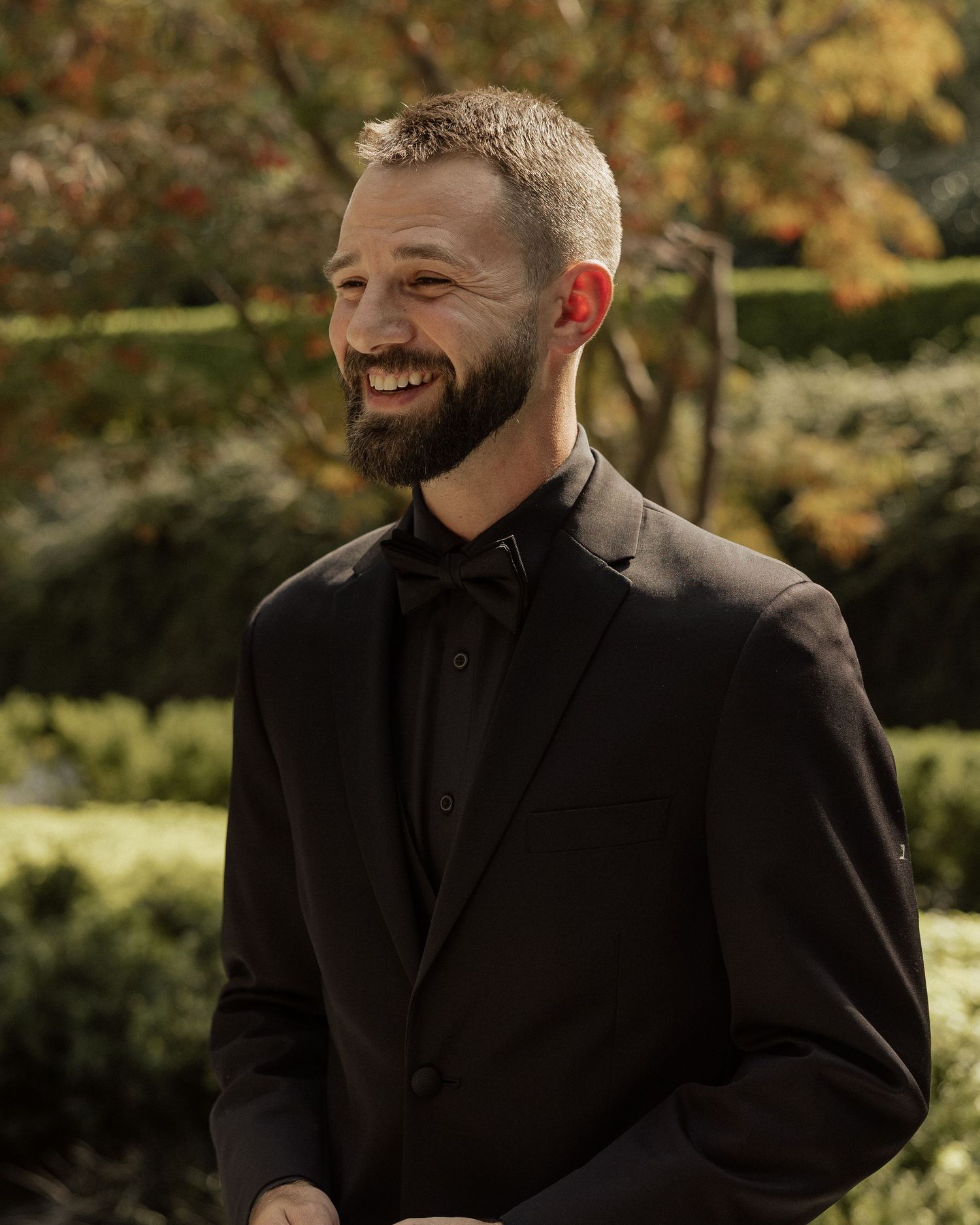 Smiling bearded man in black suit and bow tie standing outdoors in a garden setting.