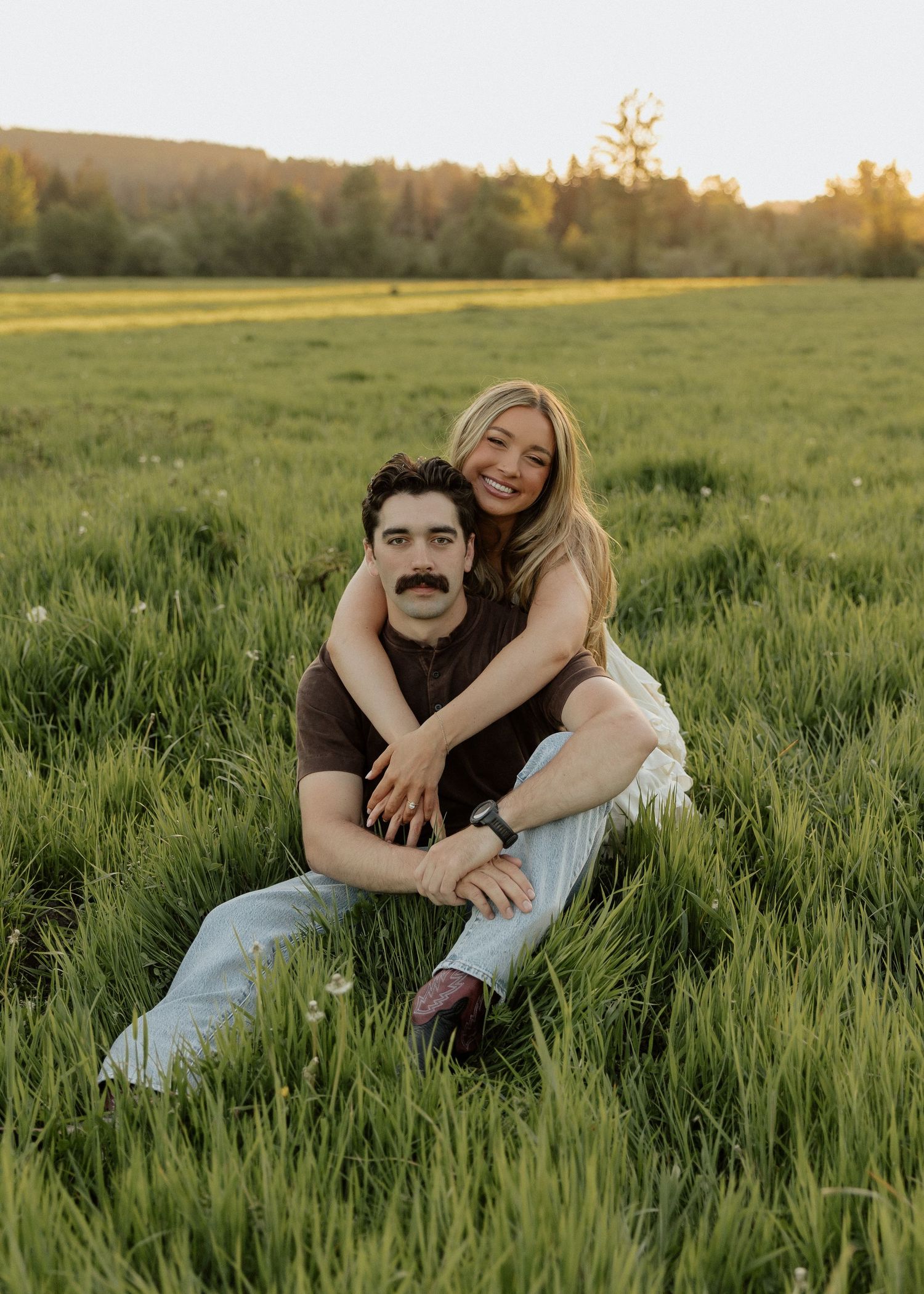 Smiling couple posing in a sunlit green field, woman embracing man from behind.