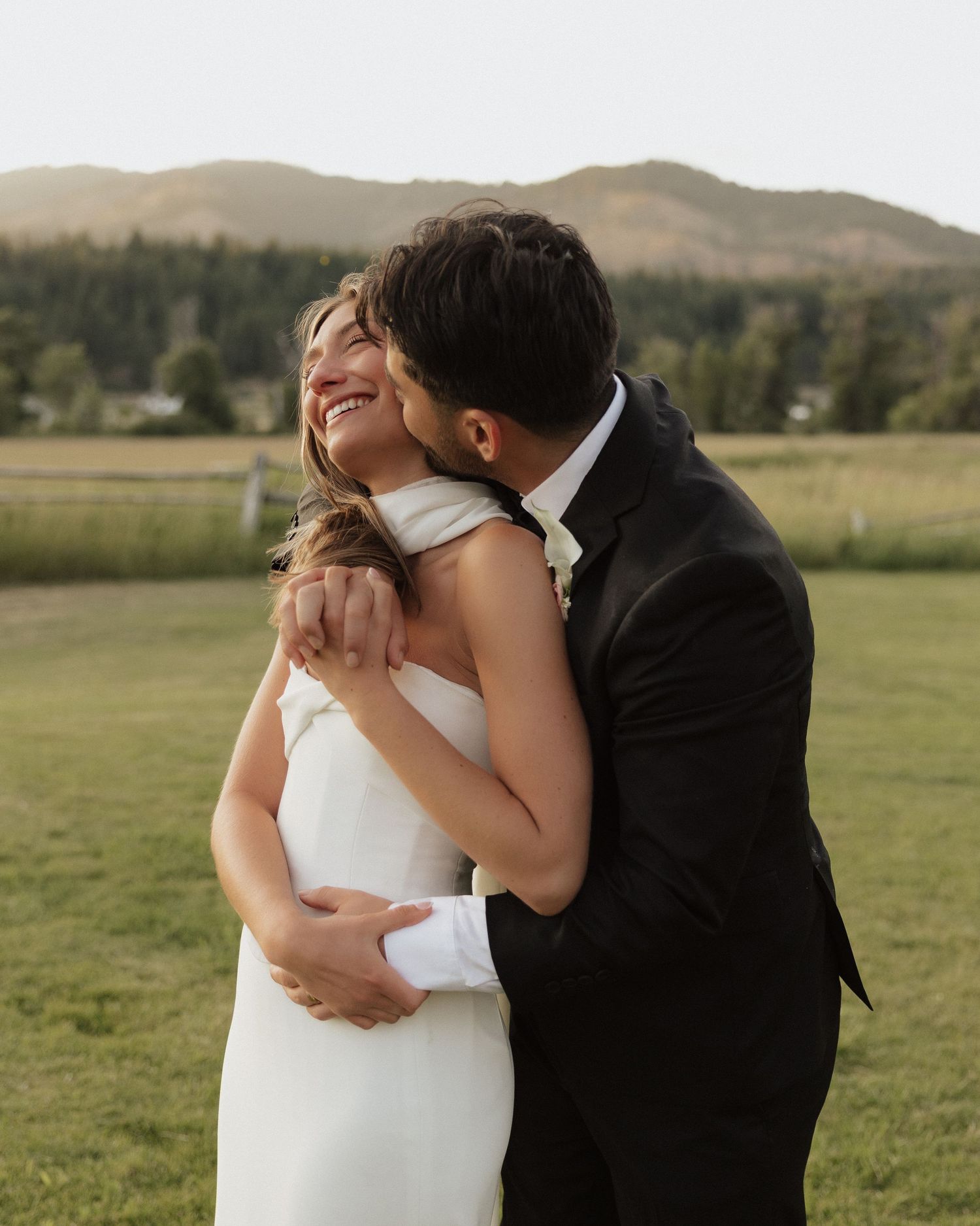 A joyful bride in a white gown laughs as her groom in a black suit embraces her outdoors with mountains behind.