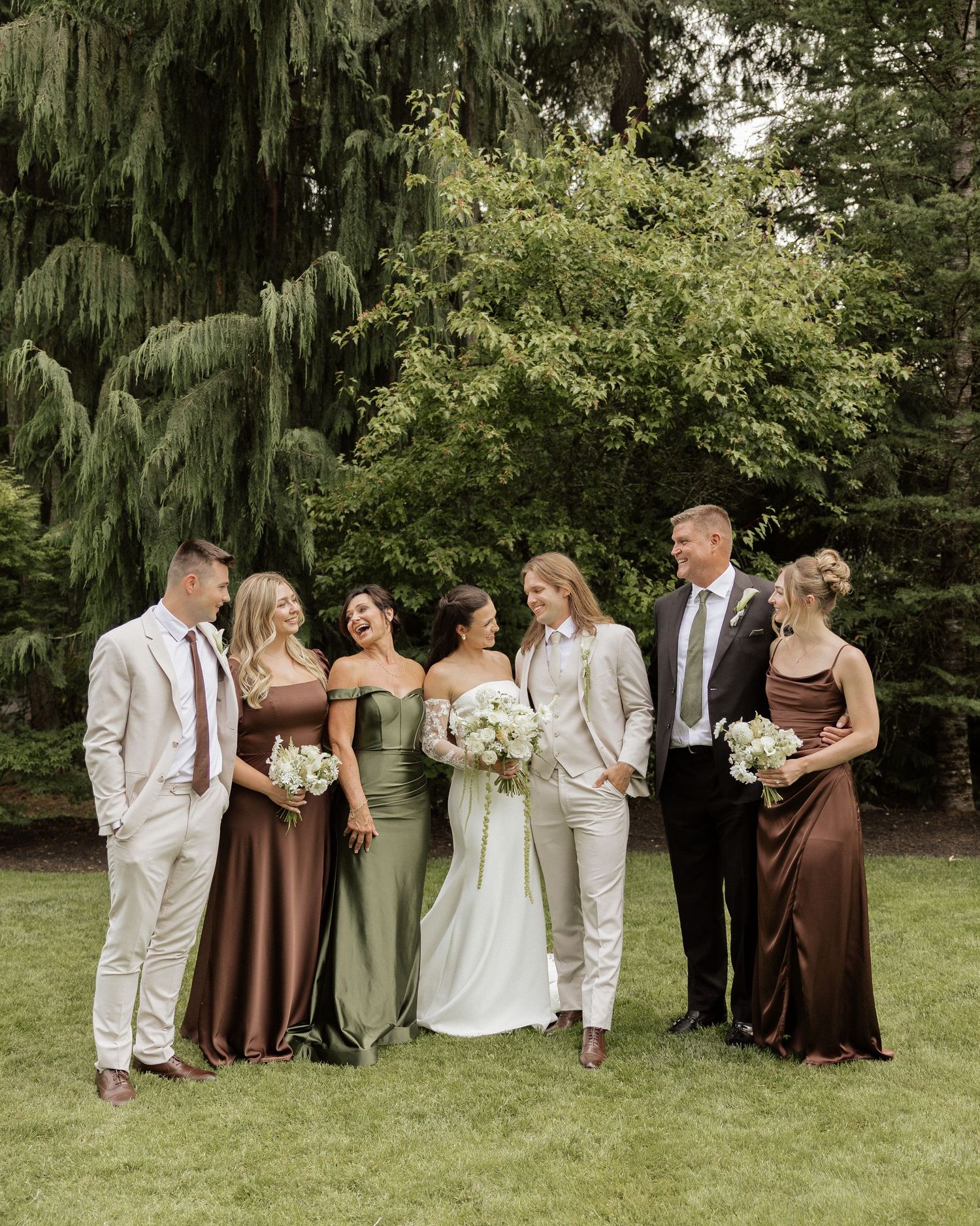 Wedding party posing outdoors in brown satin bridesmaid dresses and neutral suits with lush green trees in background.