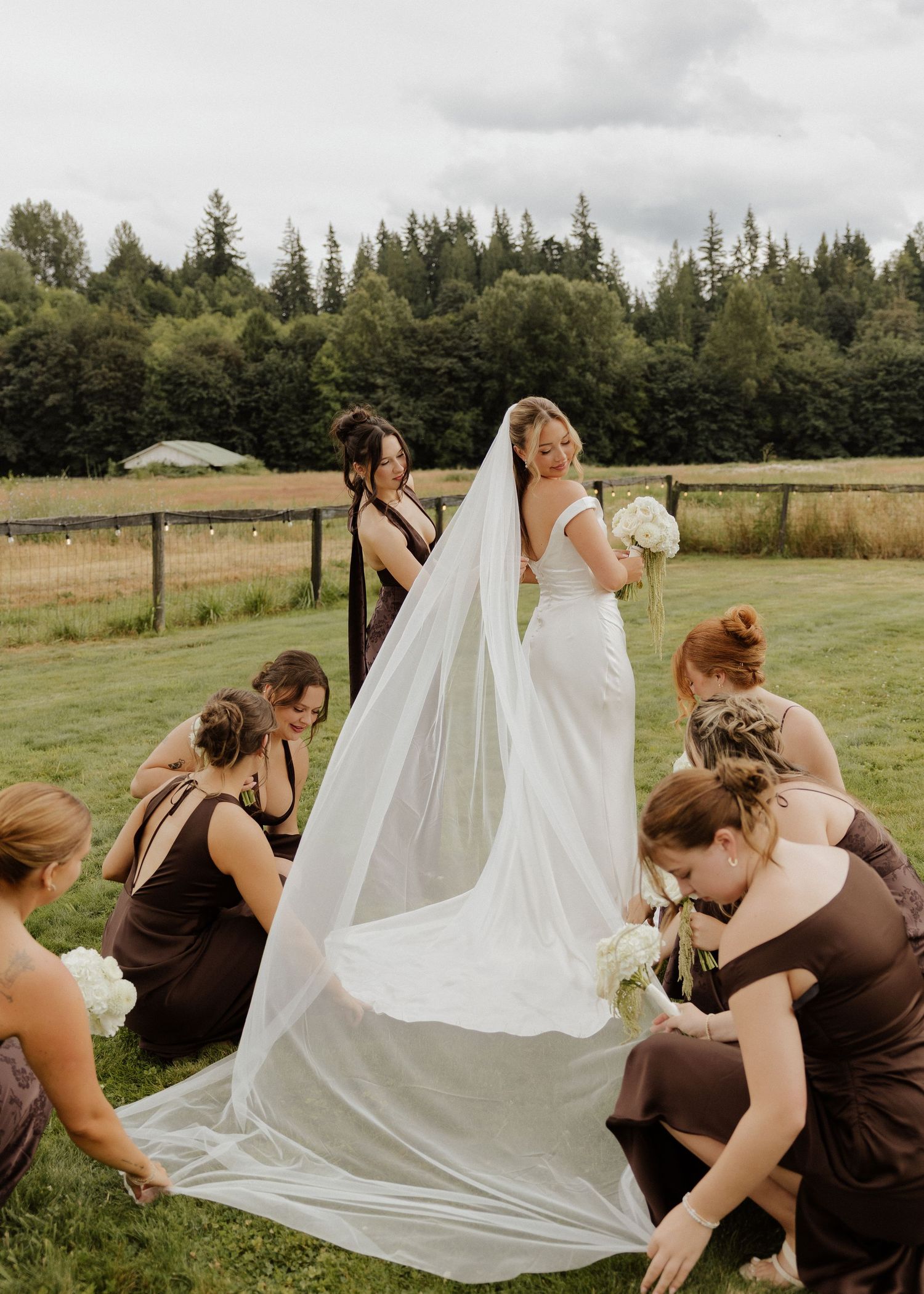 Bridesmaids in brown dresses arranging a bride's long cathedral veil on a green lawn at an outdoor wedding.