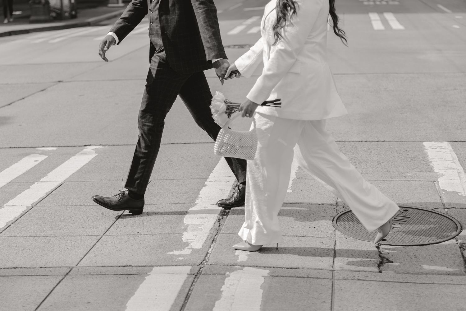 Black and white photo of a couple holding hands crossing a street crosswalk, wearing formal wedding attire.