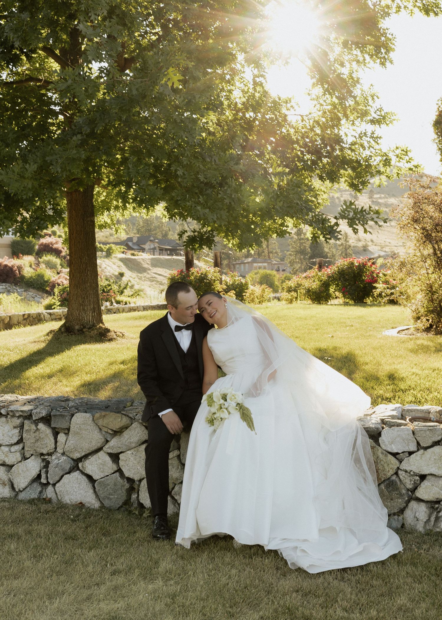 Bride and groom share romantic moment by stone wall with golden sunset light filtering through trees.