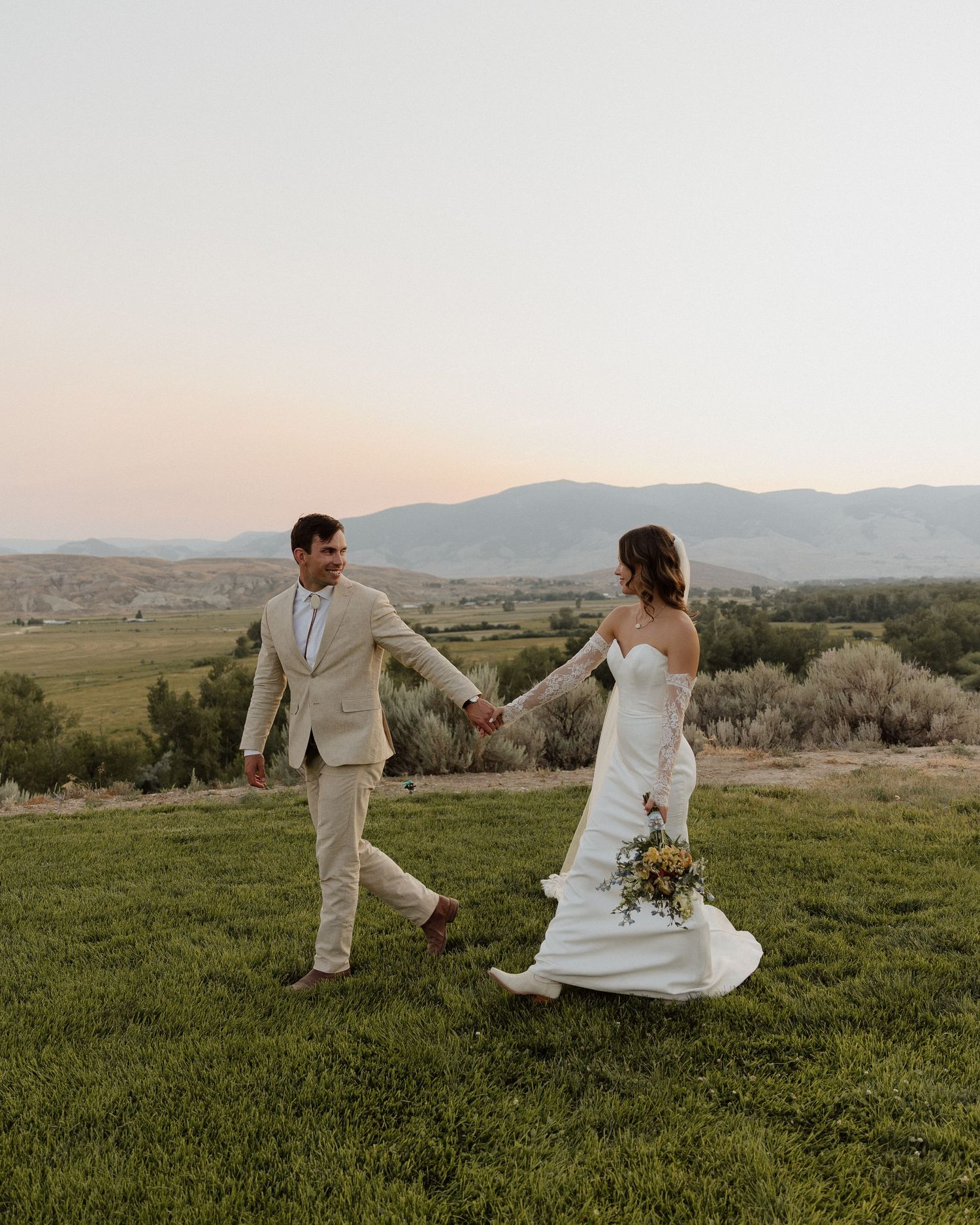 Wedding couple holds hands while walking playfully through mountain meadow at sunset.