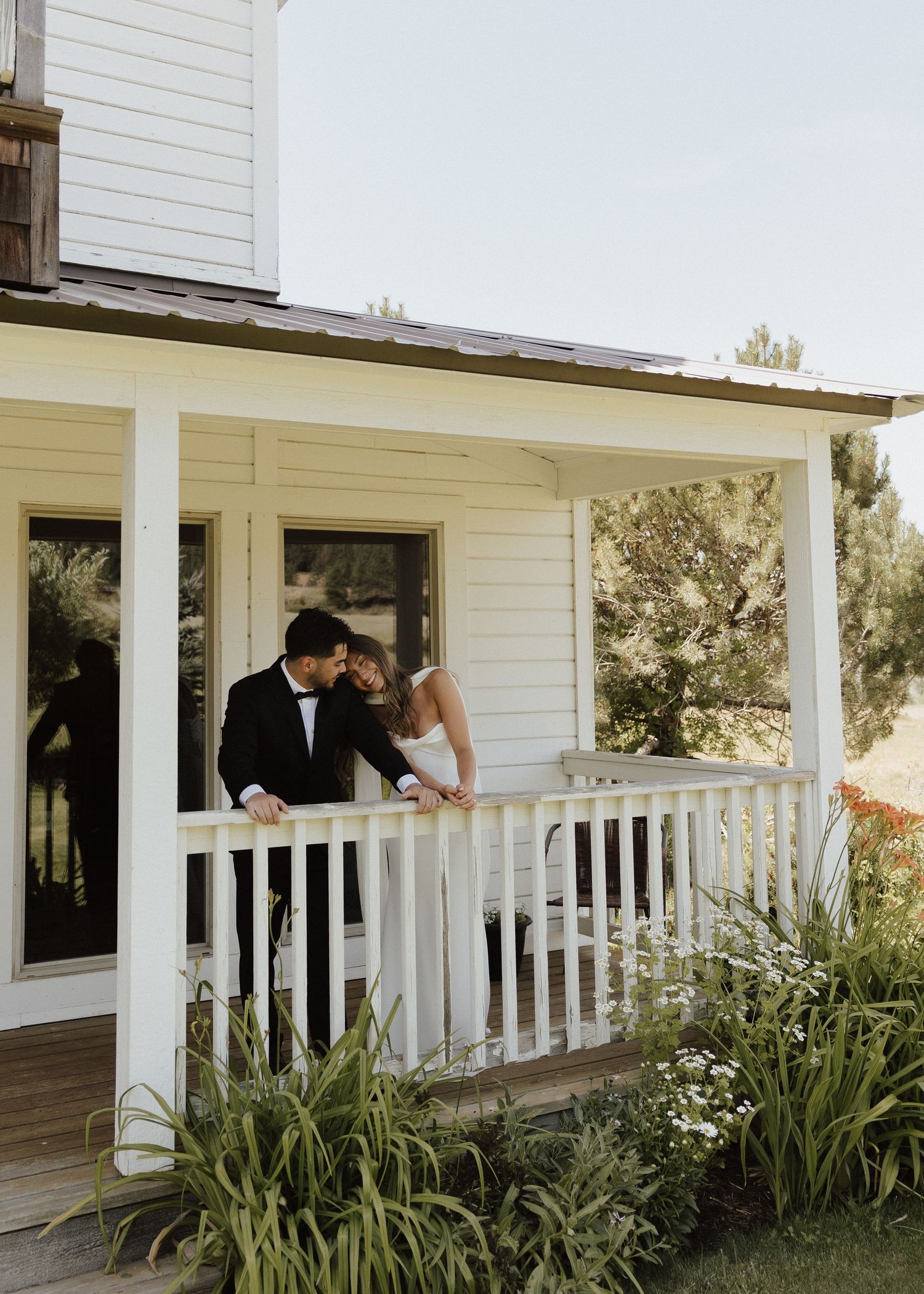 Couple sharing an intimate moment on a white farmhouse porch with white railing and decorative plants.