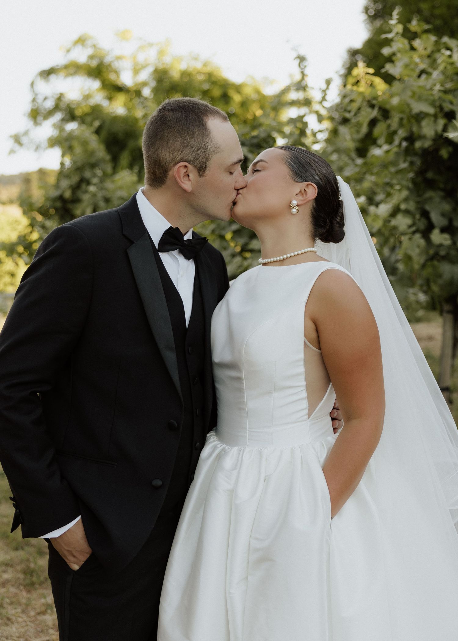 Bride and groom sharing a kiss outdoors at a vineyard, wearing a white gown and black tuxedo with bow tie.