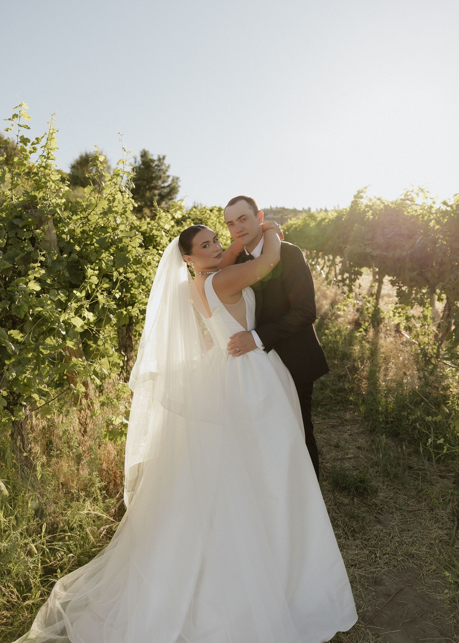 Bride in elegant white gown and groom in black suit embrace in a sunlit vineyard setting.