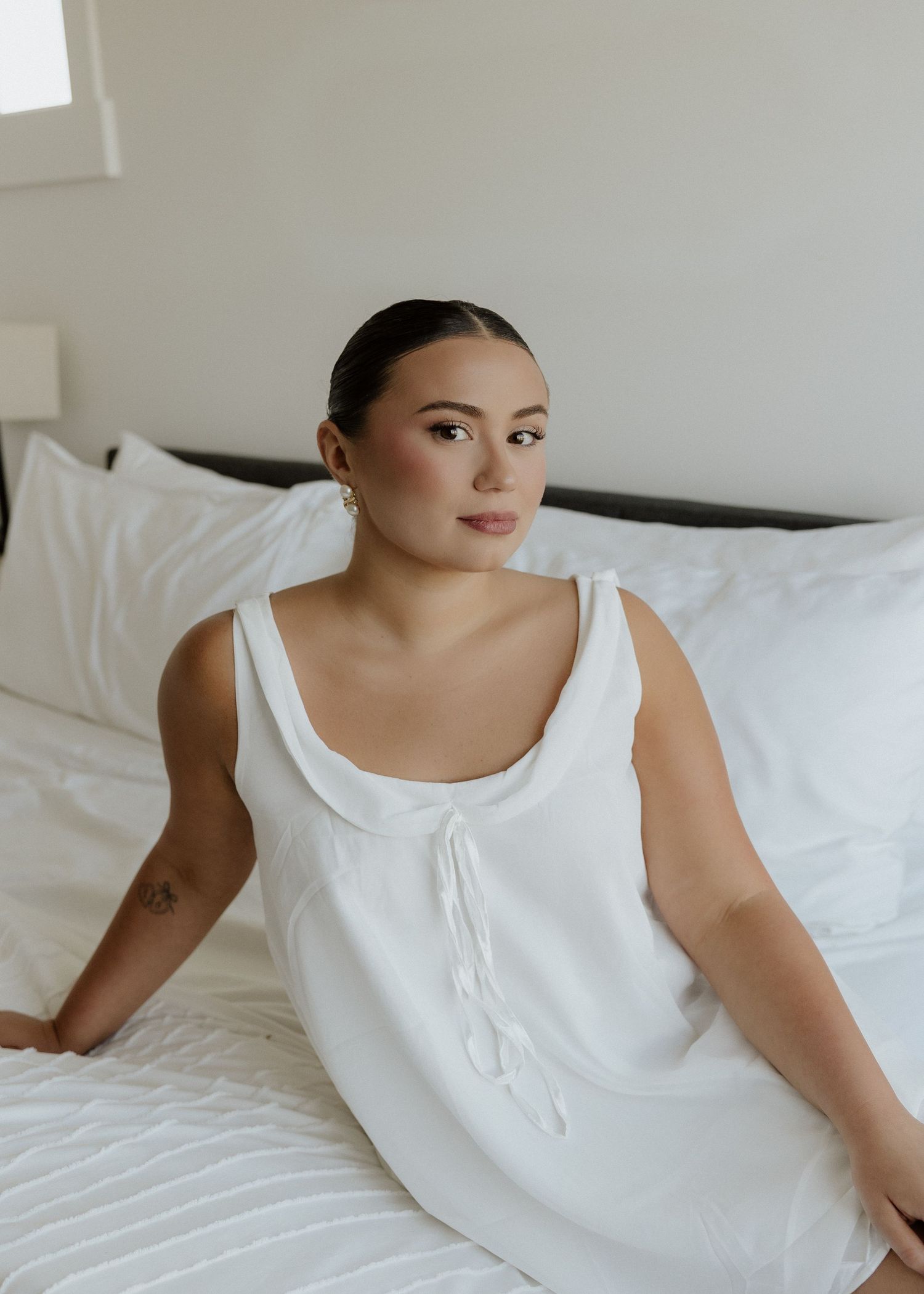 Woman in white sleeveless dress sitting on white bed with pillows, minimalist bedroom setting.
