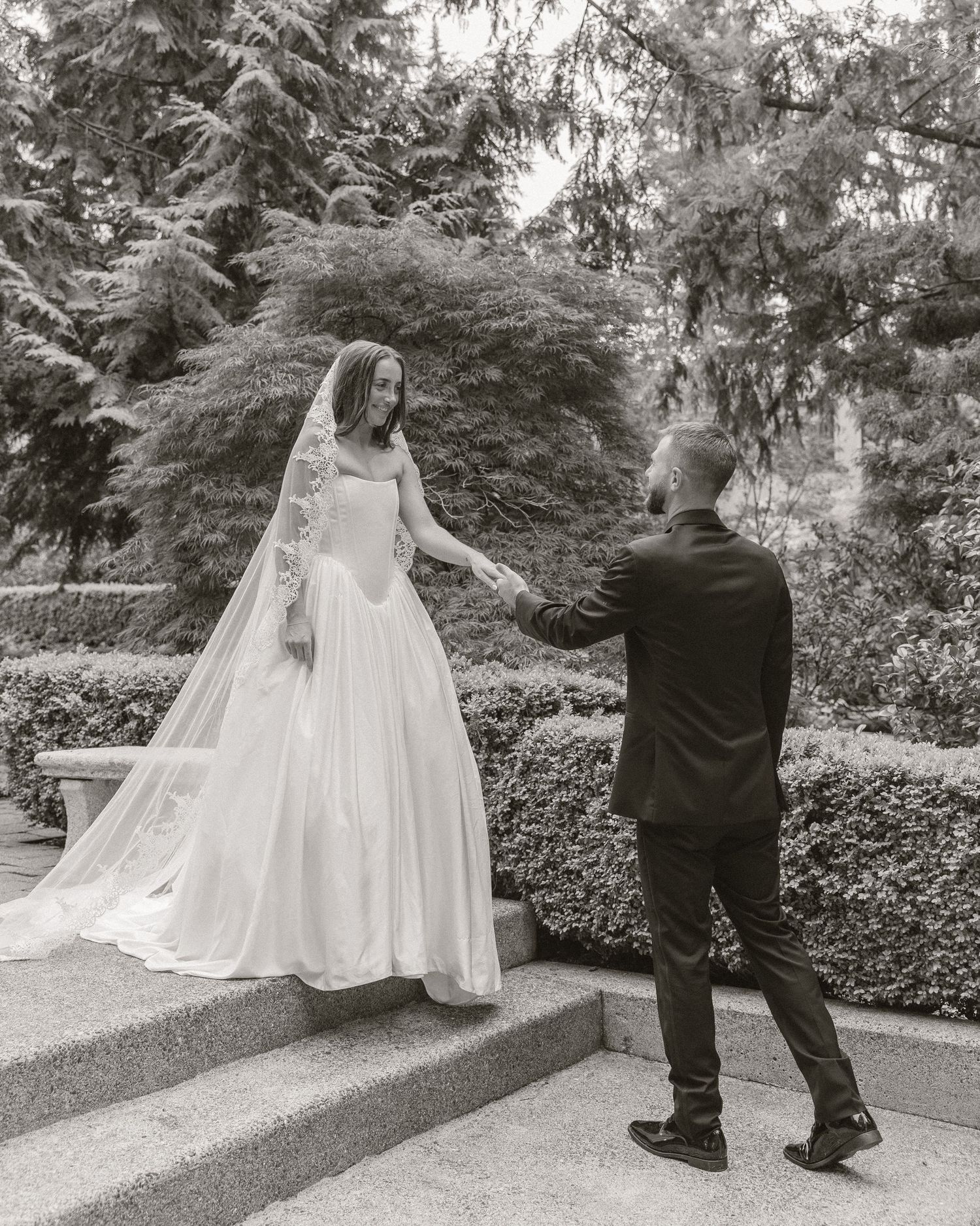 Black and white photo of a bride in a flowing gown and veil holding hands with a groom on stone steps in a garden.