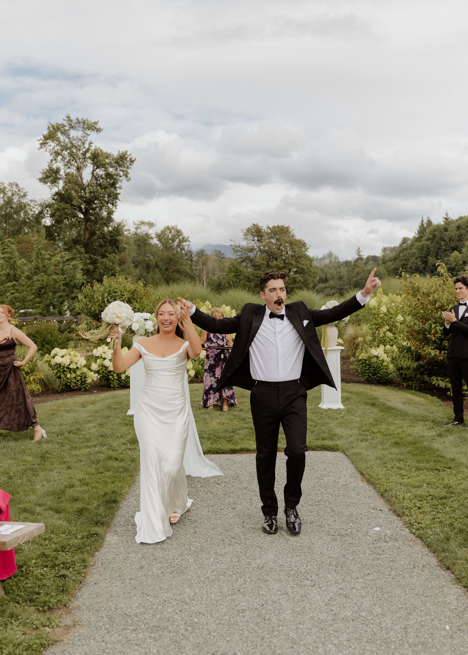 Joyful newlyweds celebrate their recessional walk down an outdoor garden pathway surrounded by guests.