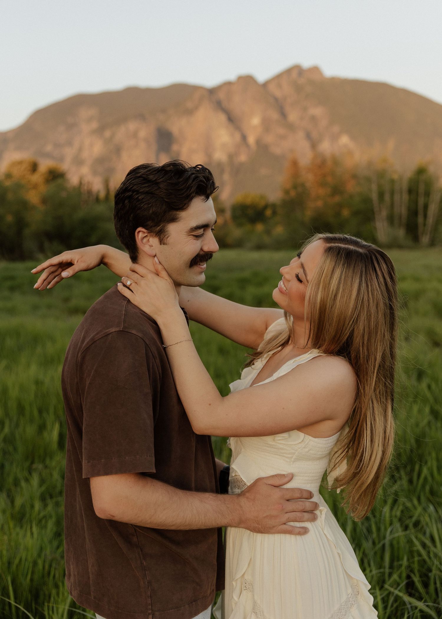 Couple embracing in a green field with a mountain sunset backdrop, woman in white dress smiling at man.