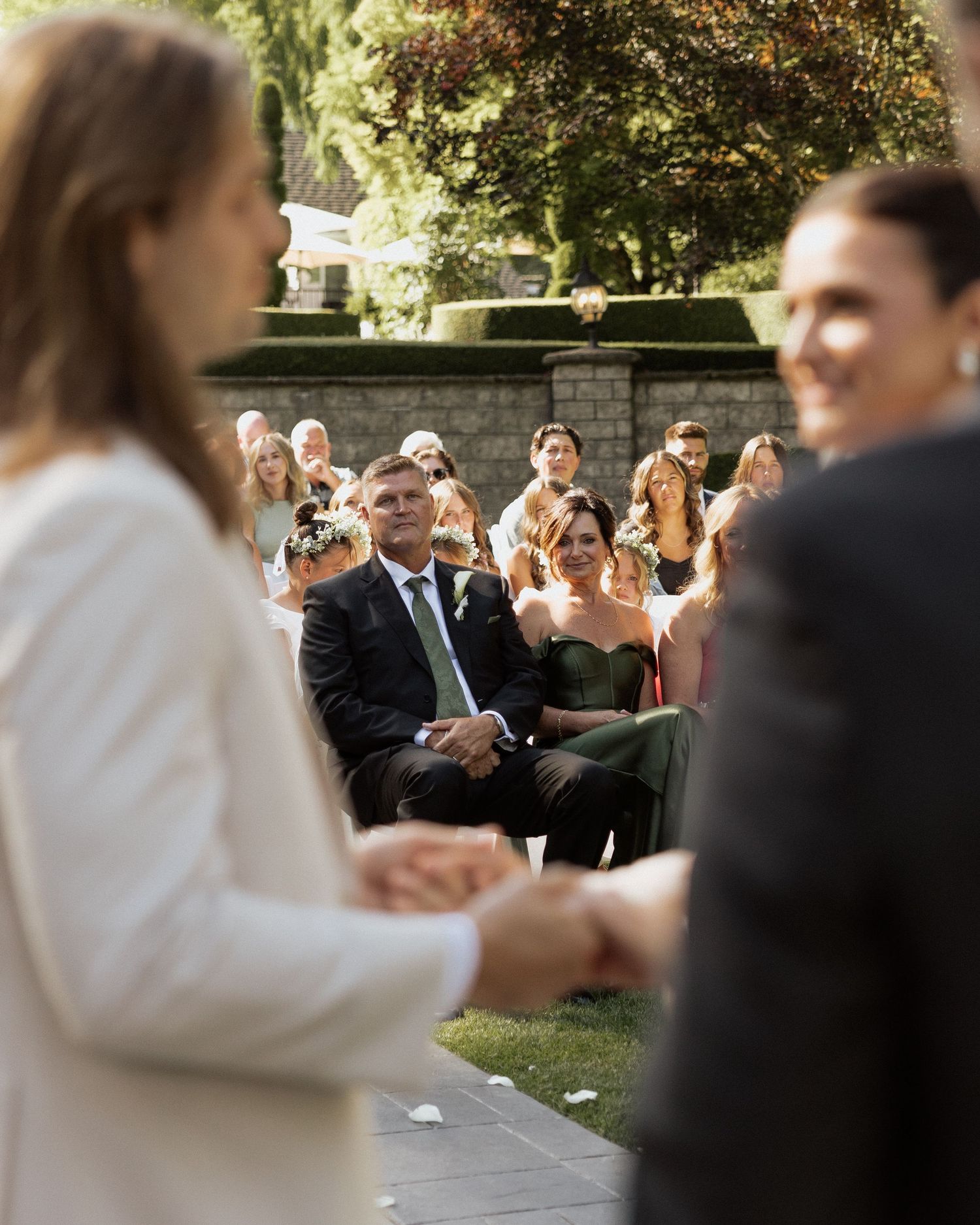 Wedding guests smile and applaud as couple holds hands during an outdoor garden ceremony.
