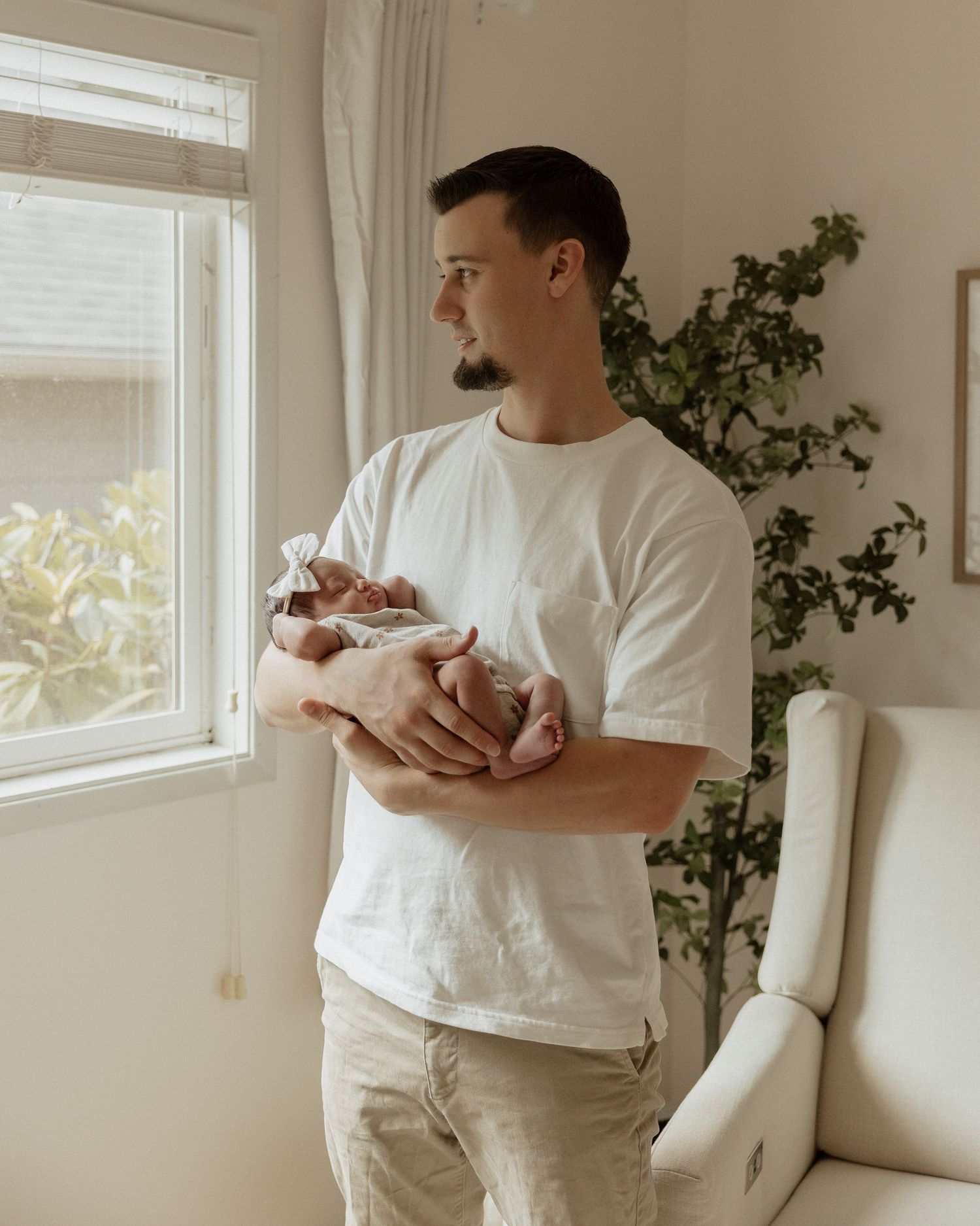 Parent standing by window holding newborn baby while gazing outside in natural light.