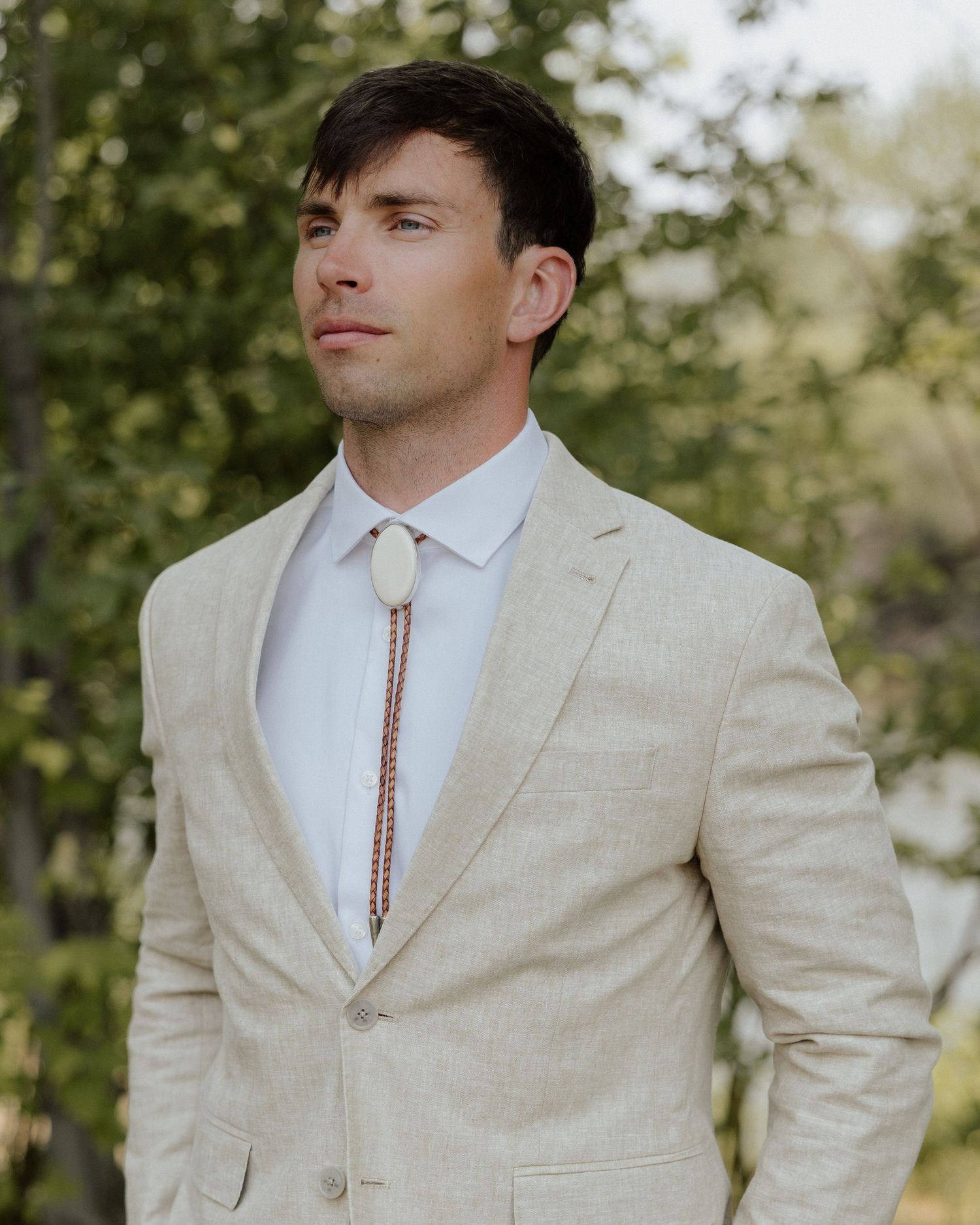 Person wearing a beige suit with a bolo tie against a natural outdoor backdrop.