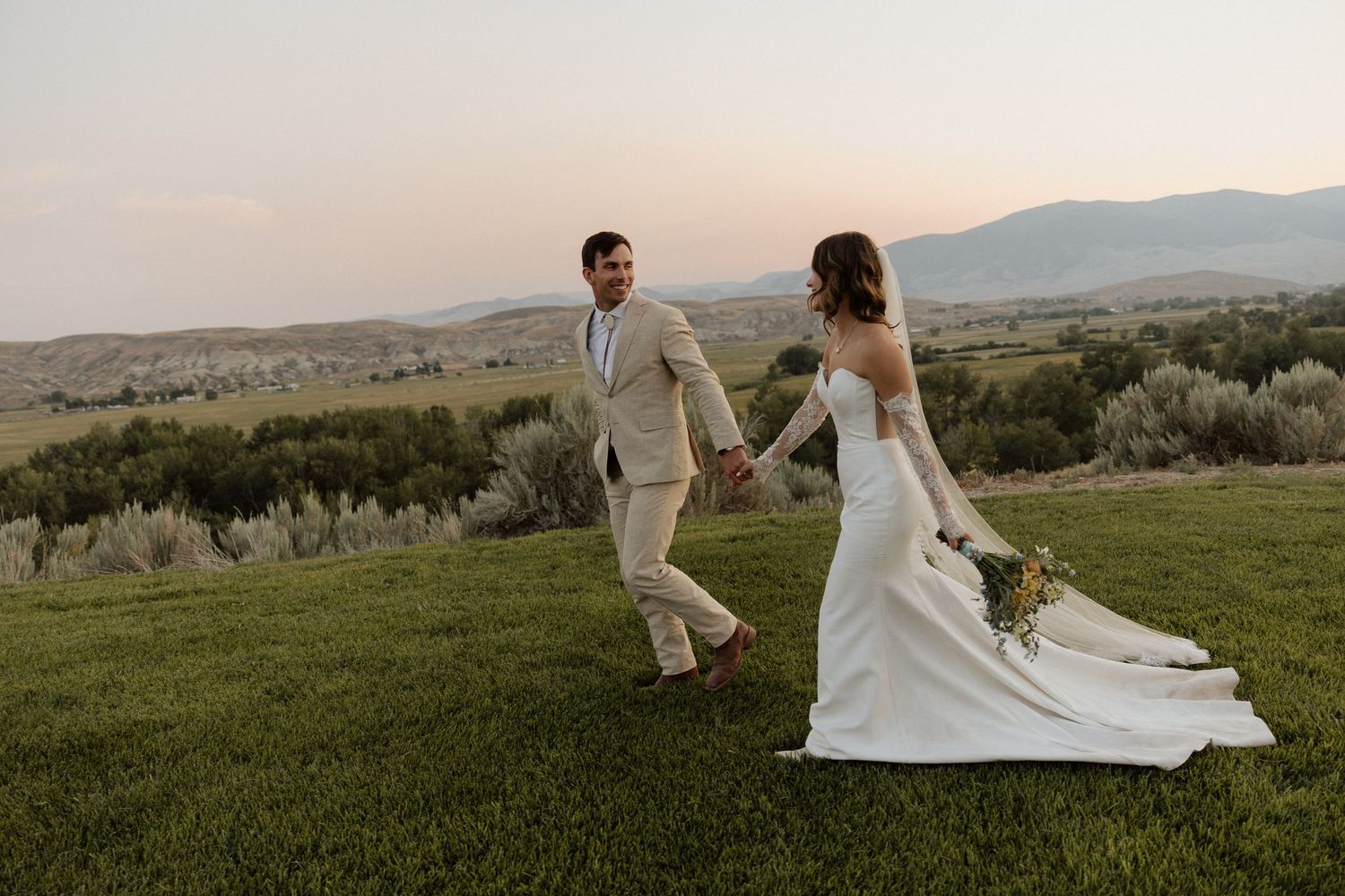 Couple walking hand in hand on hilltop during sunset in wedding attire.
