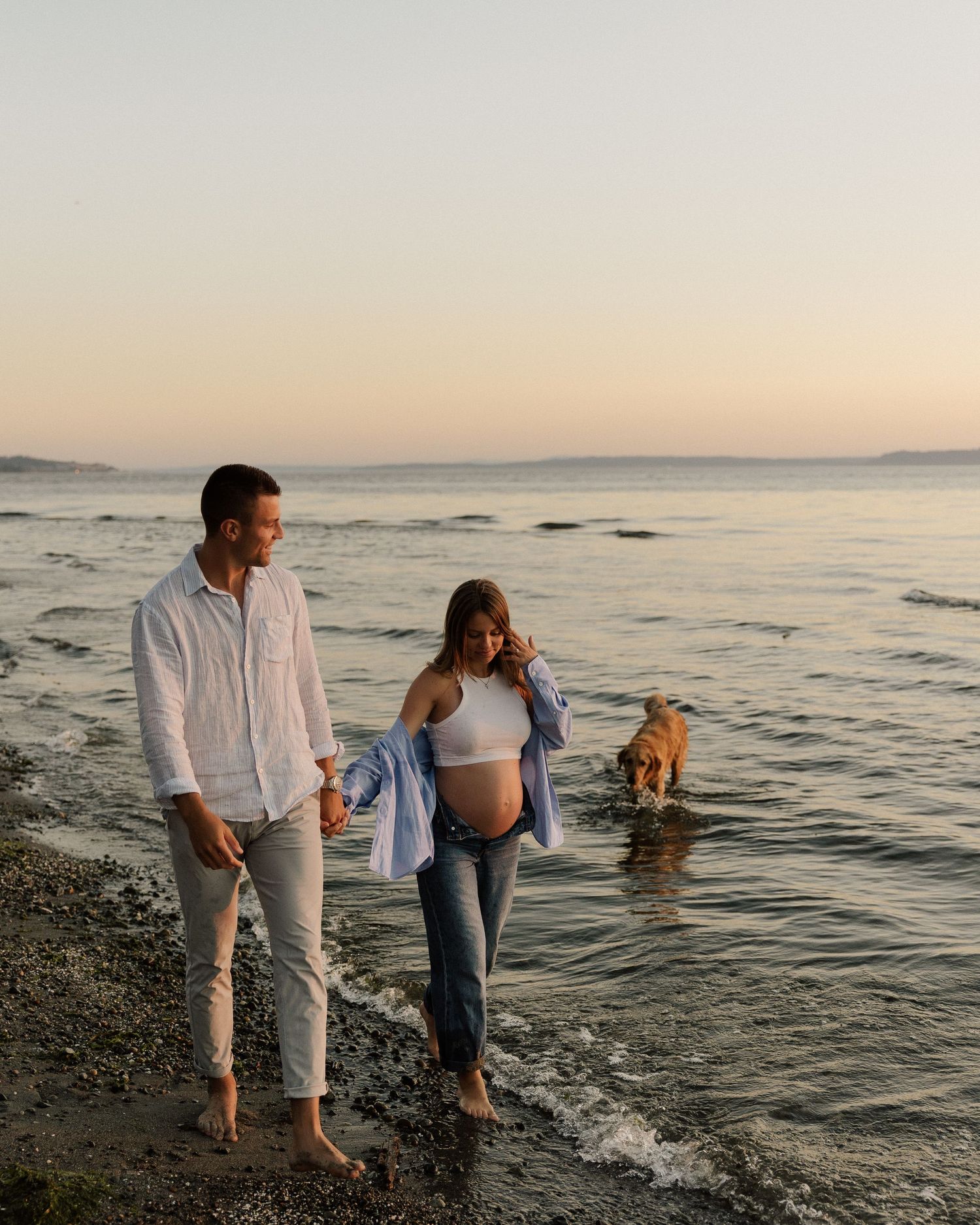 Couple walking barefoot along beach with their dog during sunset maternity session.
