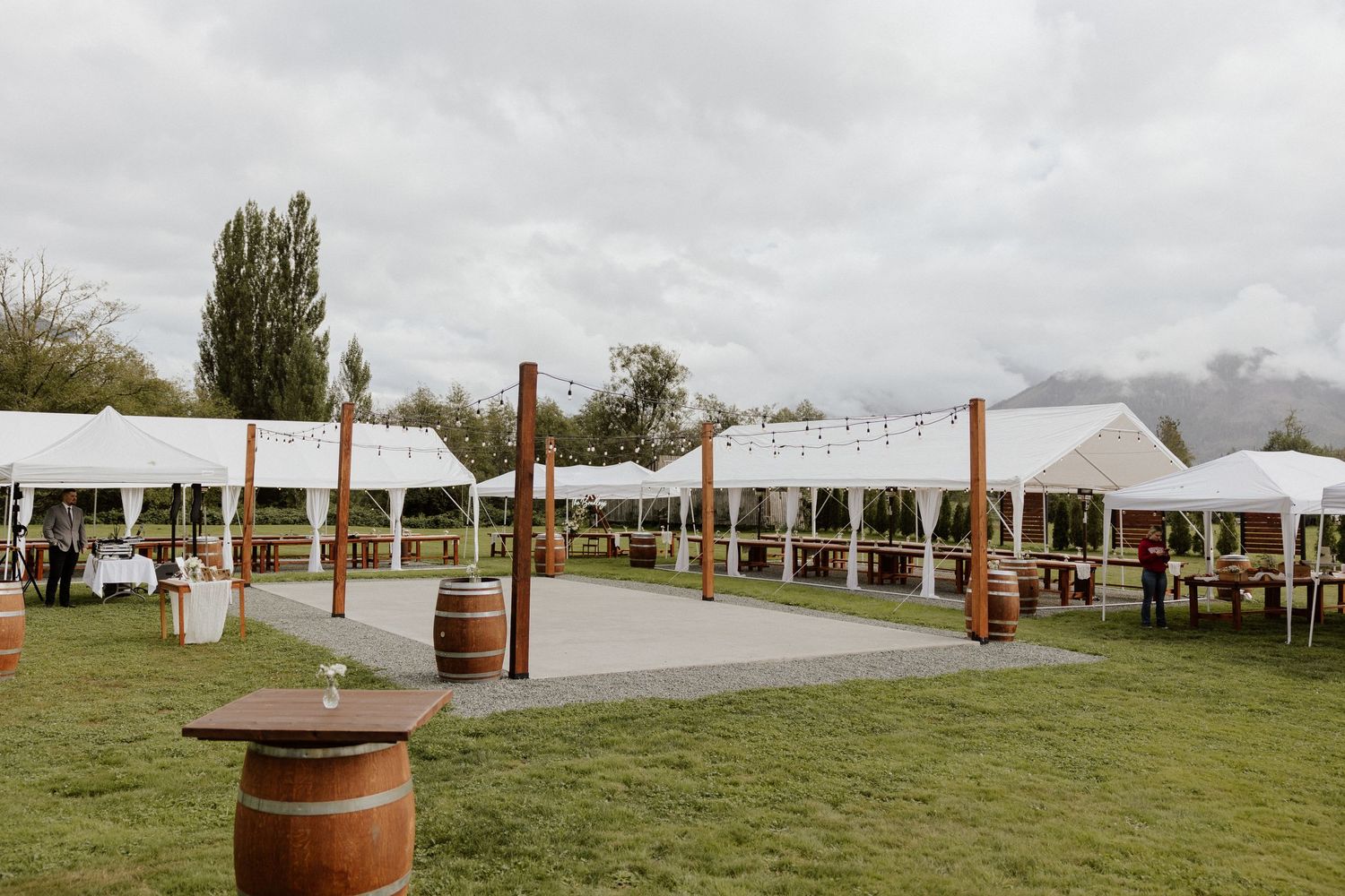 Outdoor wedding reception area with white tents and wooden barrel decor on grass.