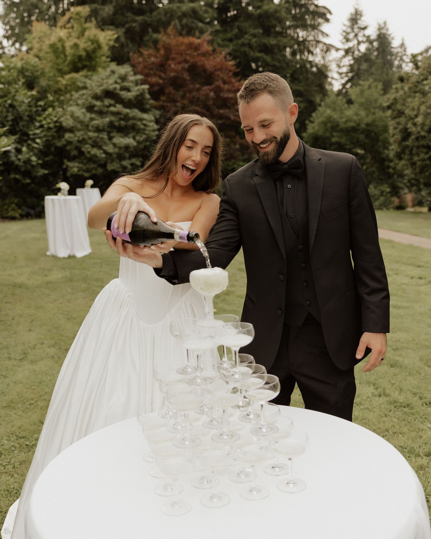 Smiling bride and groom pouring champagne tower at outdoor wedding reception.