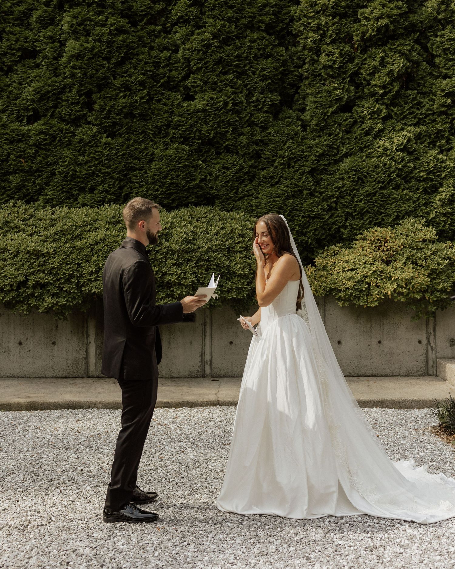 Bride in white gown and groom in black suit exchange vows outdoors with green hedge backdrop.