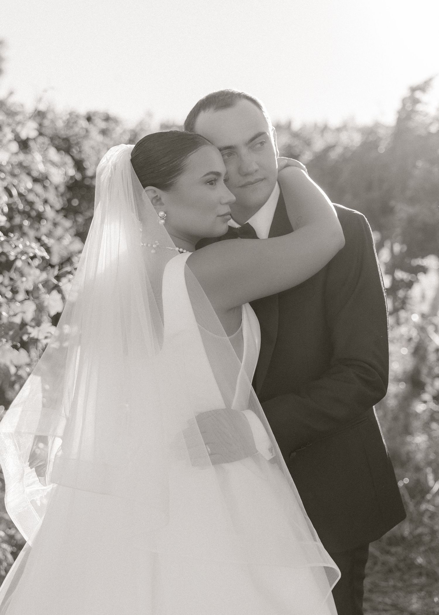 Black and white wedding photo of bride in veil embracing groom in suit outdoors among lush greenery.