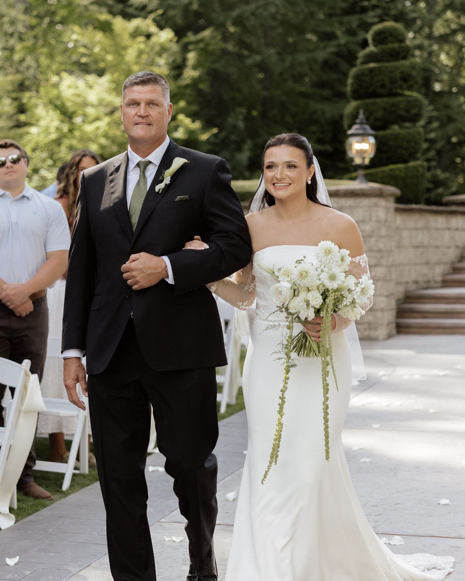 Bride in white gown holding bouquet walks down outdoor aisle with her father in black suit at elegant wedding ceremony.
