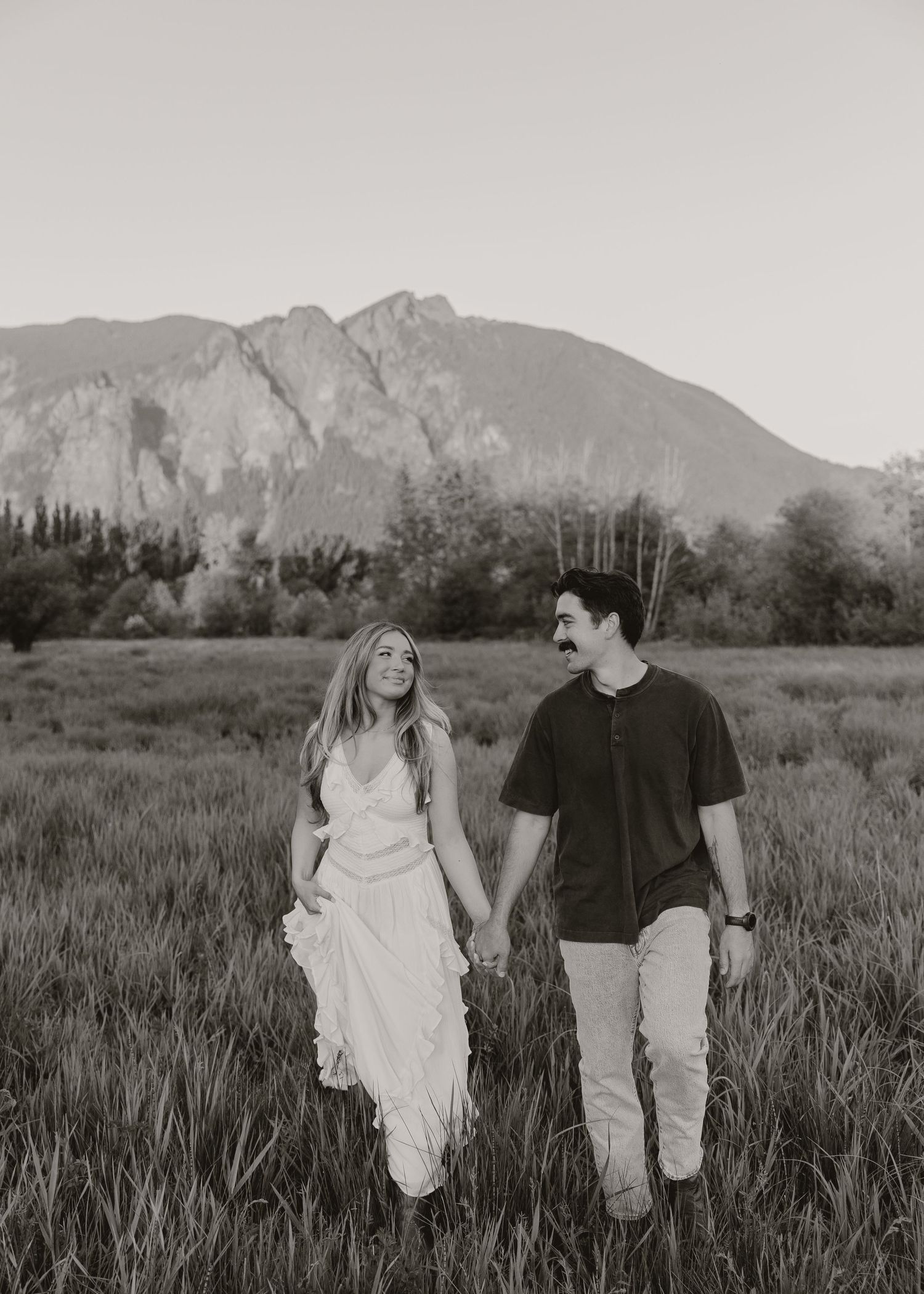 Black and white photo of couple holding hands walking in a field with mountain backdrop.