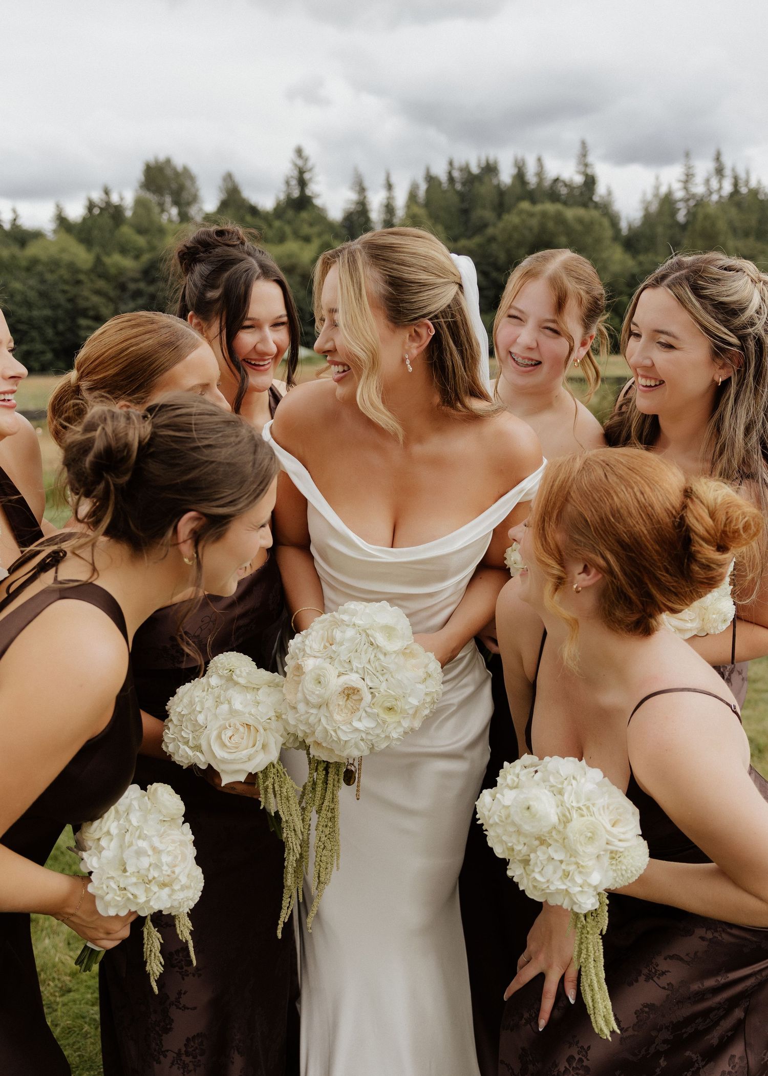 Bride in white gown surrounded by bridesmaids in dark brown dresses holding white floral bouquets outdoors.