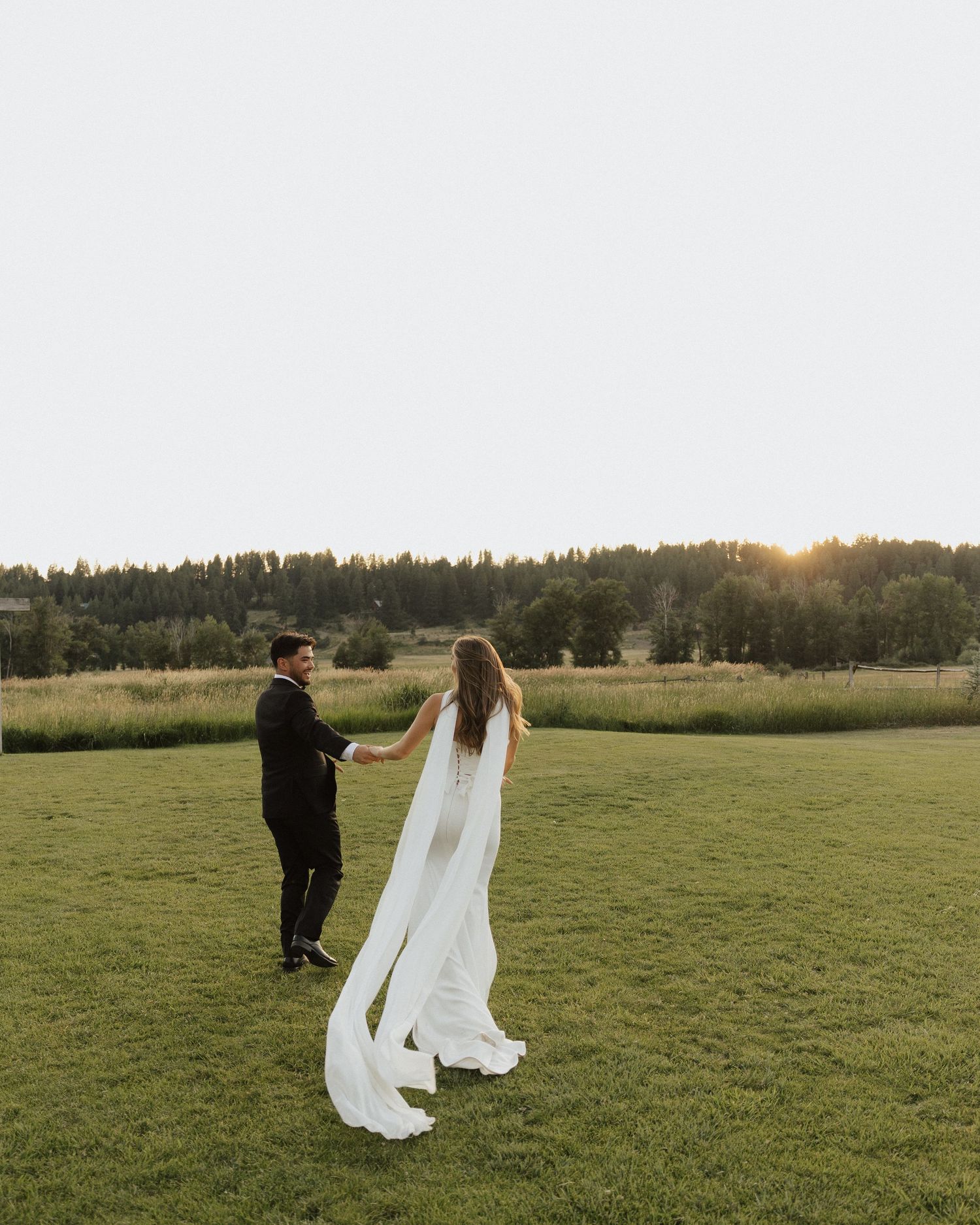 A bride in a flowing white gown and groom in black tuxedo hold hands at sunset on a lush green field.