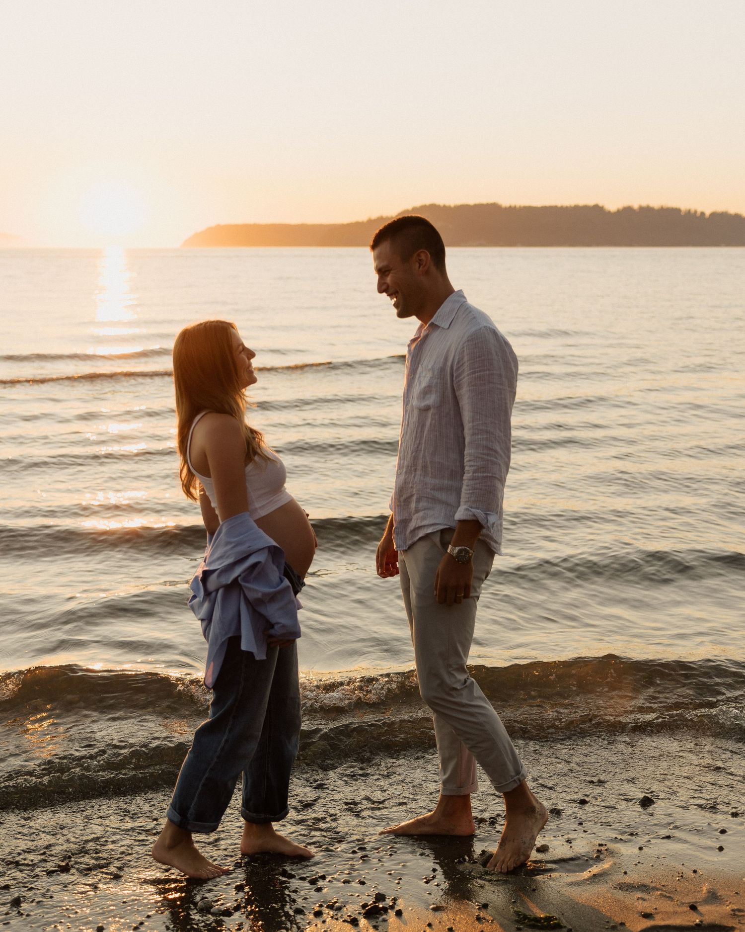 A couple facing each other on a rocky beach during golden hour with peaceful water in the background.