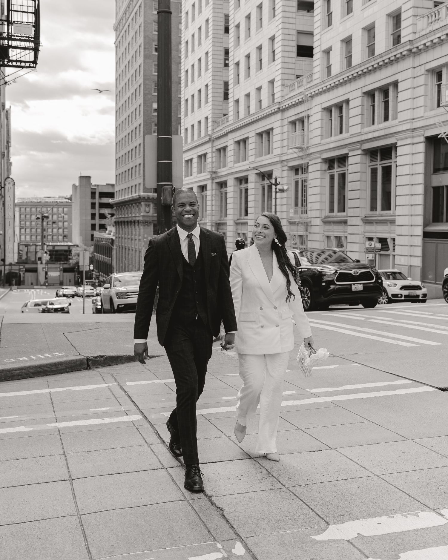 Black and white photo of a couple in formal attire crossing a city street, man in suit and woman in white blazer.