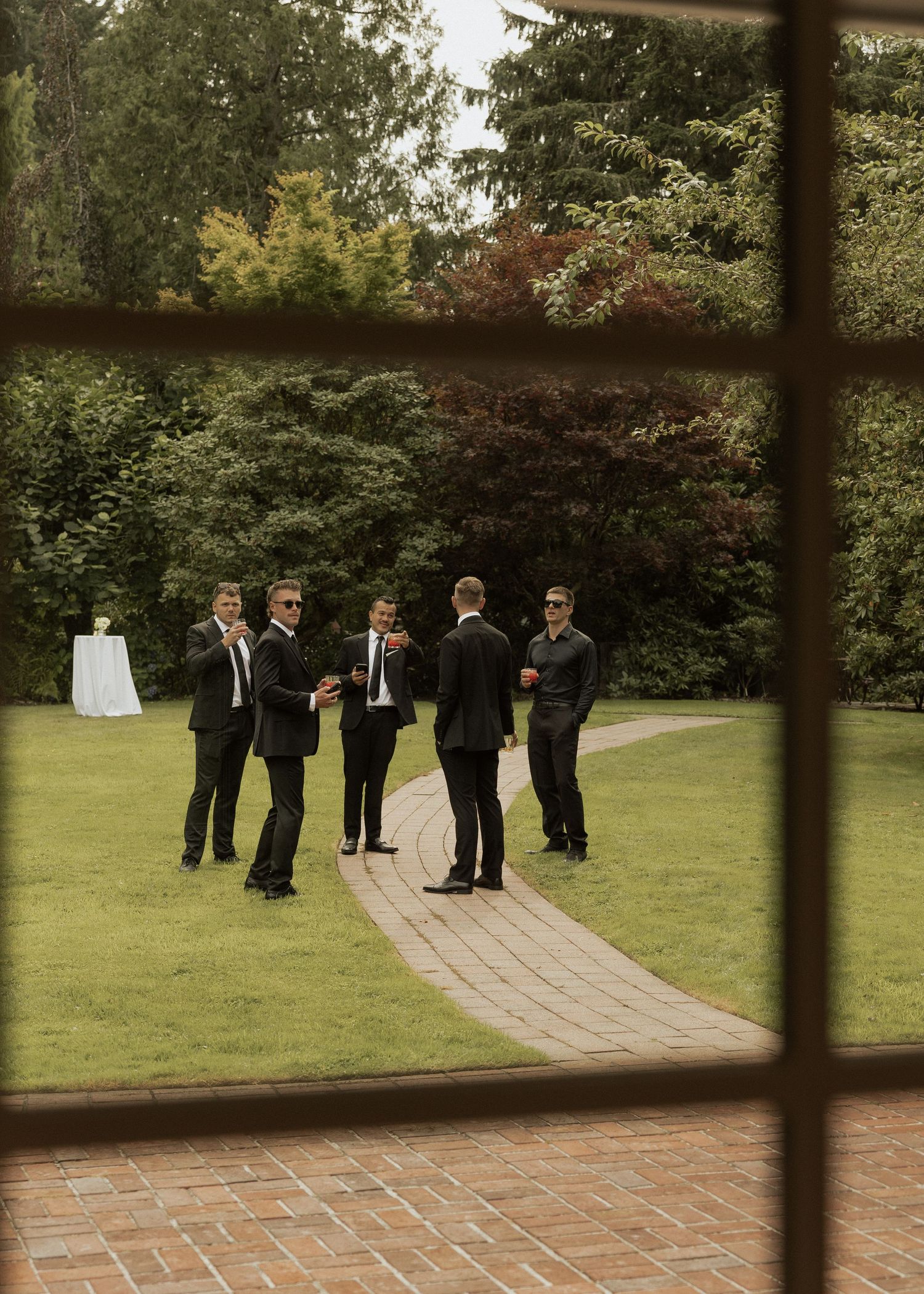 Groomsmen in black suits gathered on garden pathway, photographed through window at elegant outdoor wedding venue.