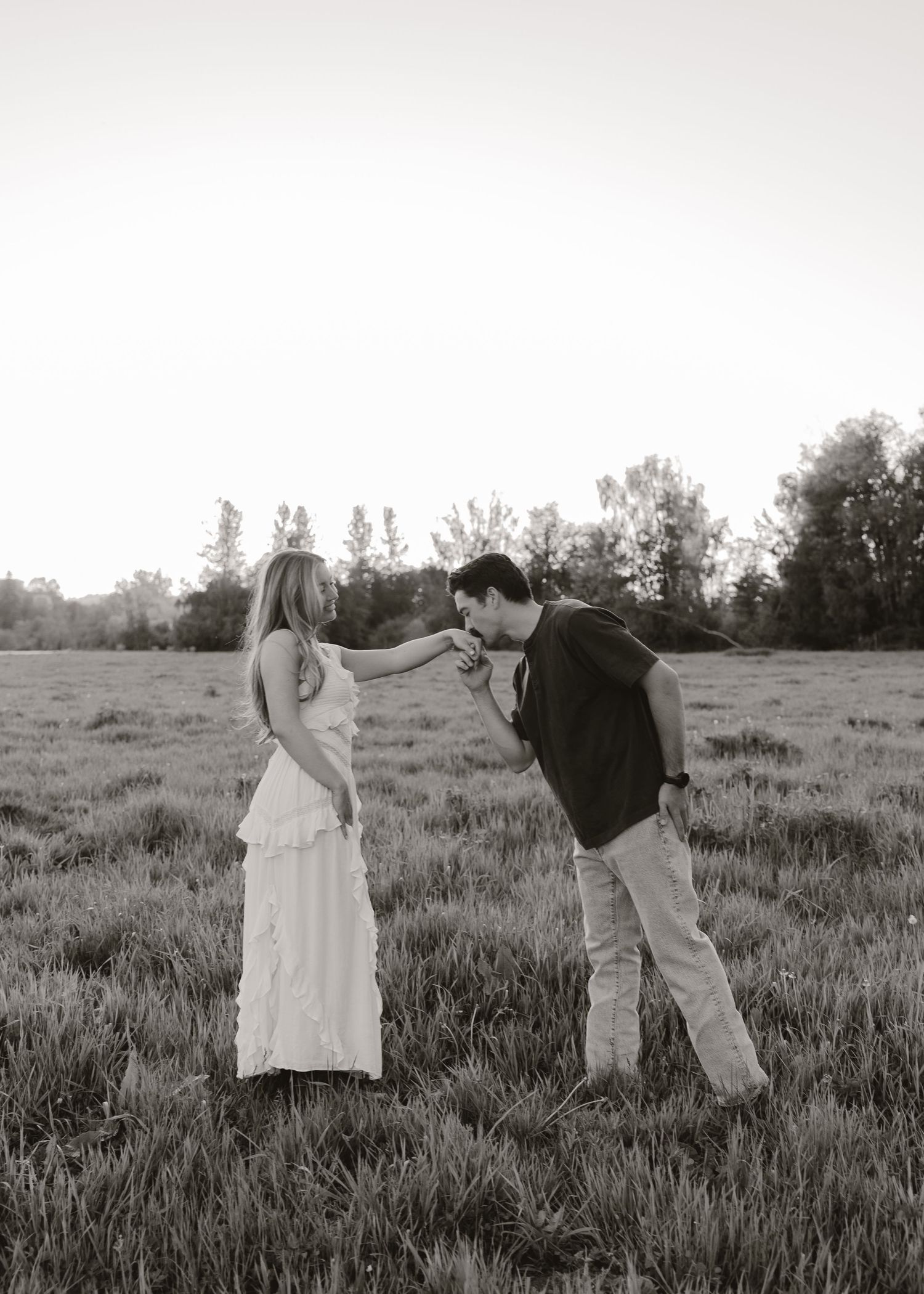 Black and white photo of a man kissing a woman's hand in a grassy field with trees in the background.