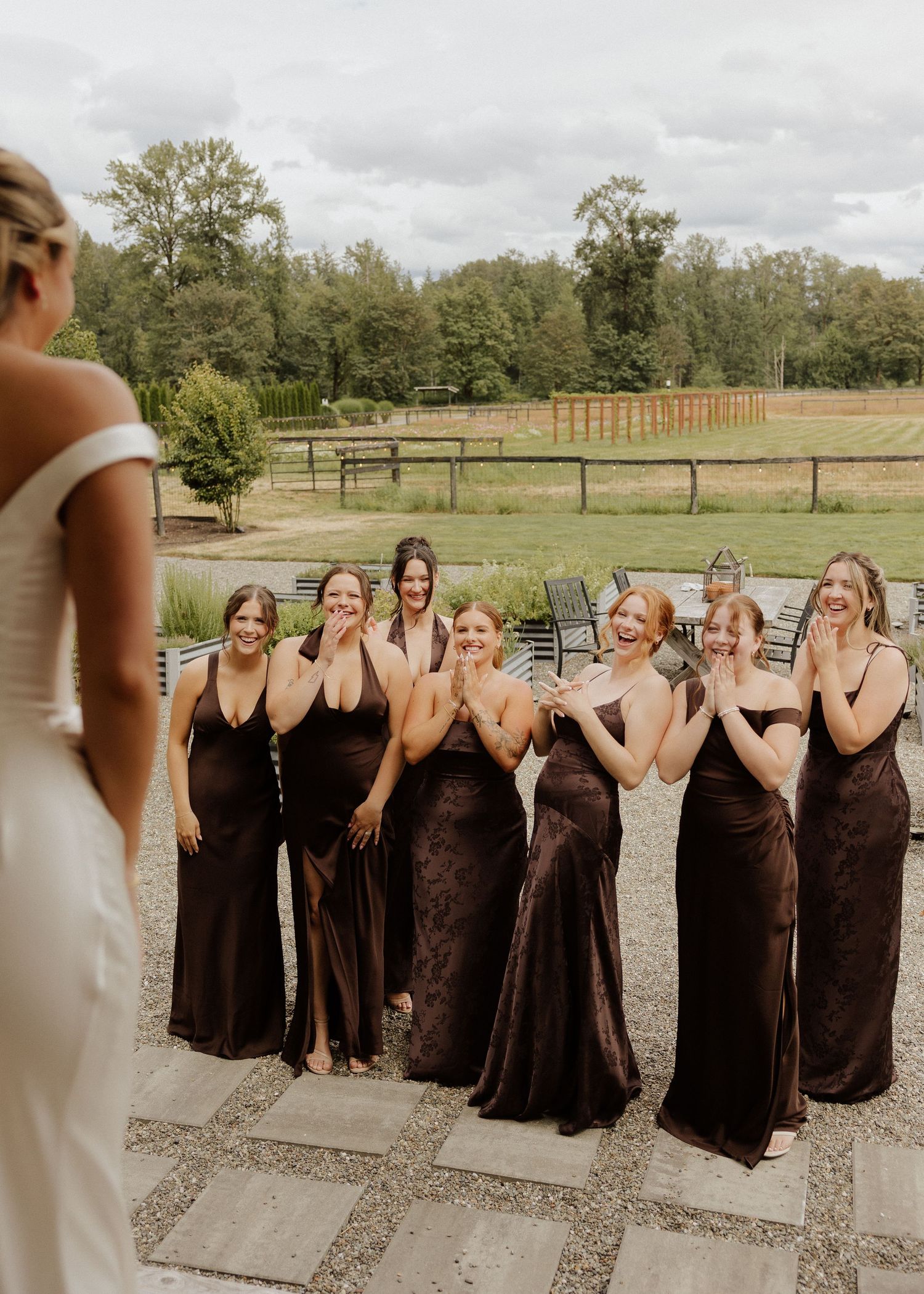 Bridesmaids in brown dresses react with joy seeing the bride in her white gown at an outdoor wedding venue.