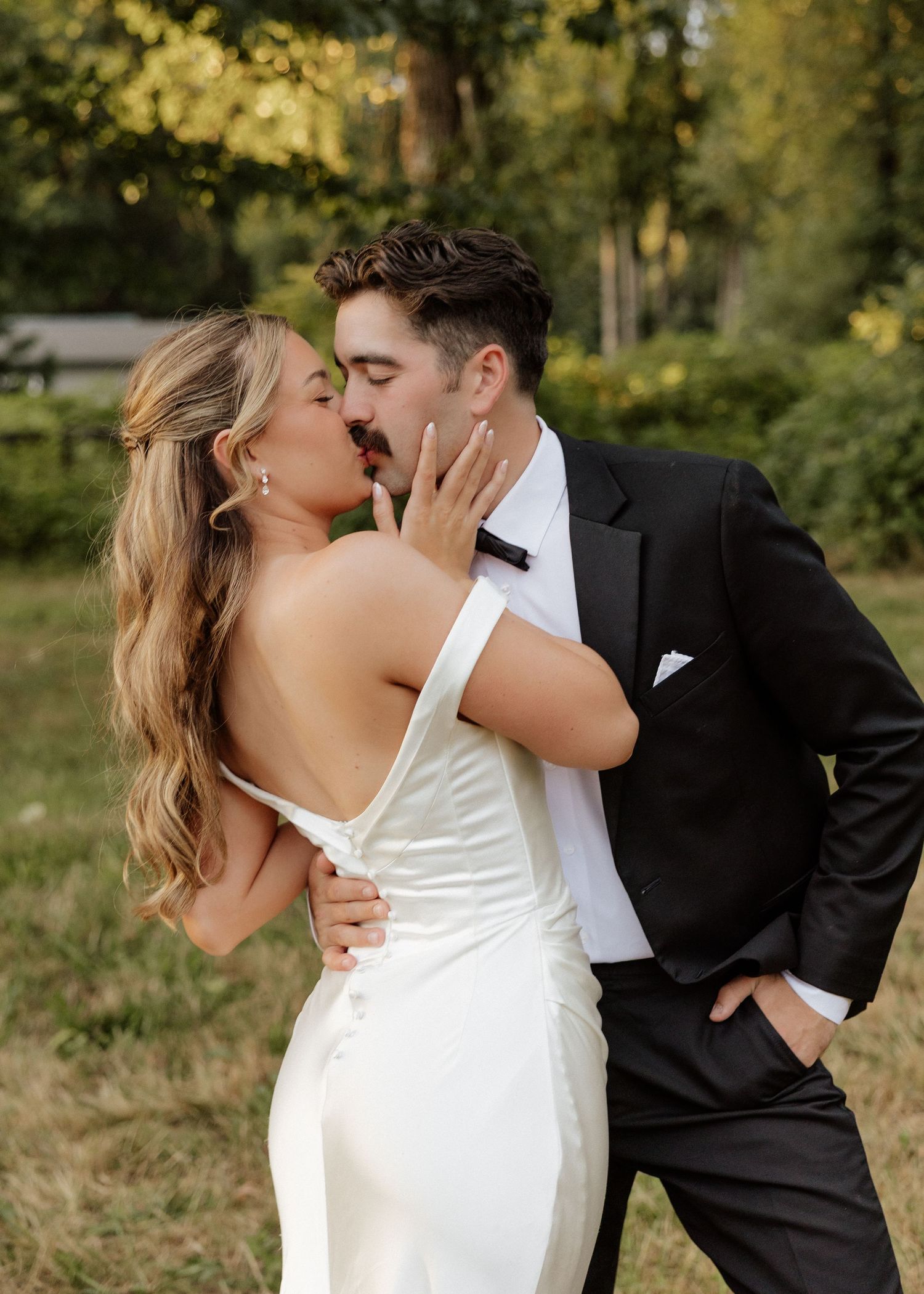 Bride and groom sharing a passionate kiss outdoors in golden light, bride in elegant backless white gown, groom in black tuxedo.